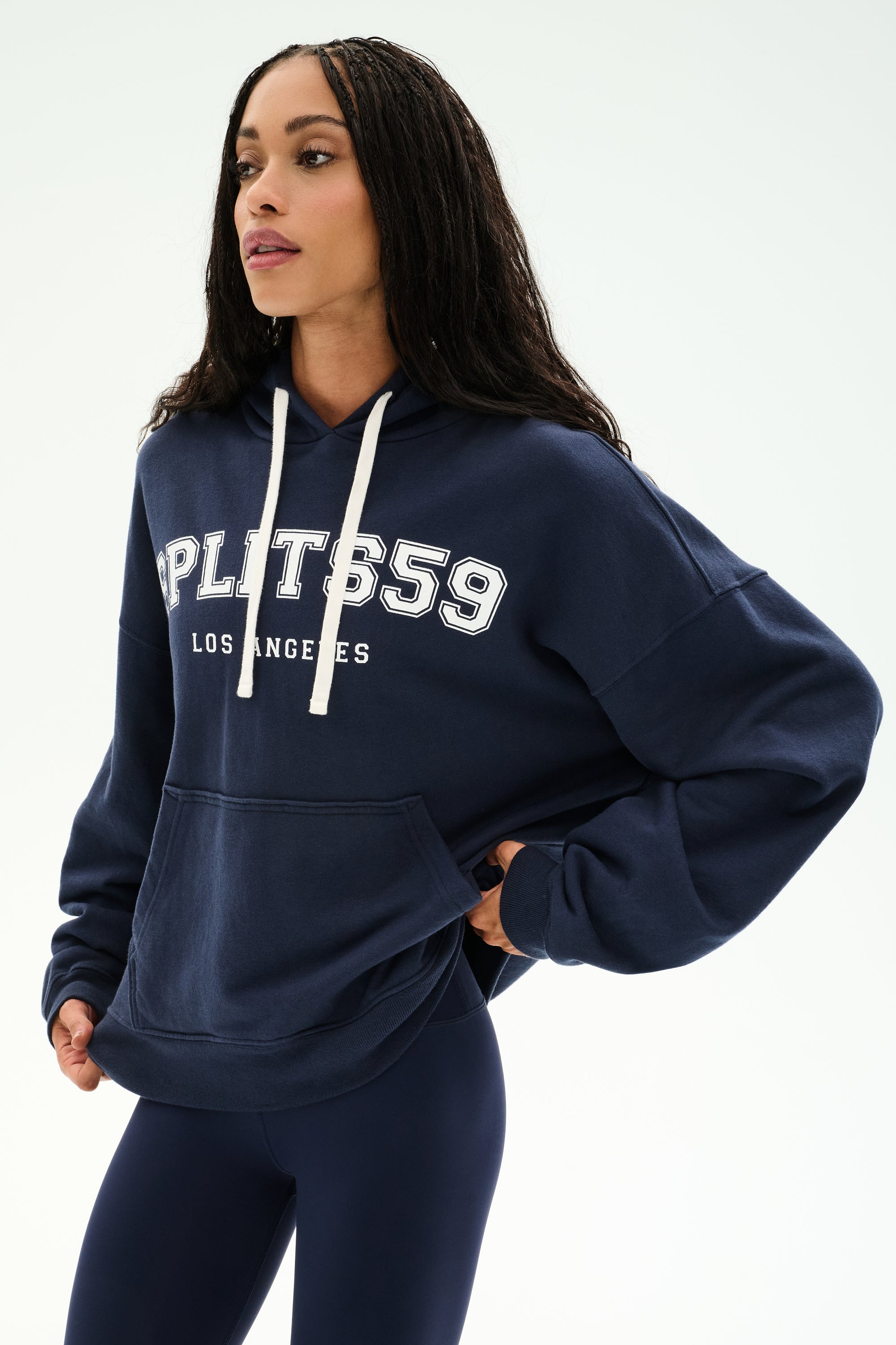 A person wearing the Stella French Terry Oversized Hoodie in indigo by SPLITS59, featuring "SPLITS59 Los Angeles" text and crafted from soft cotton fabric, paired with navy leggings while standing against a plain light background.