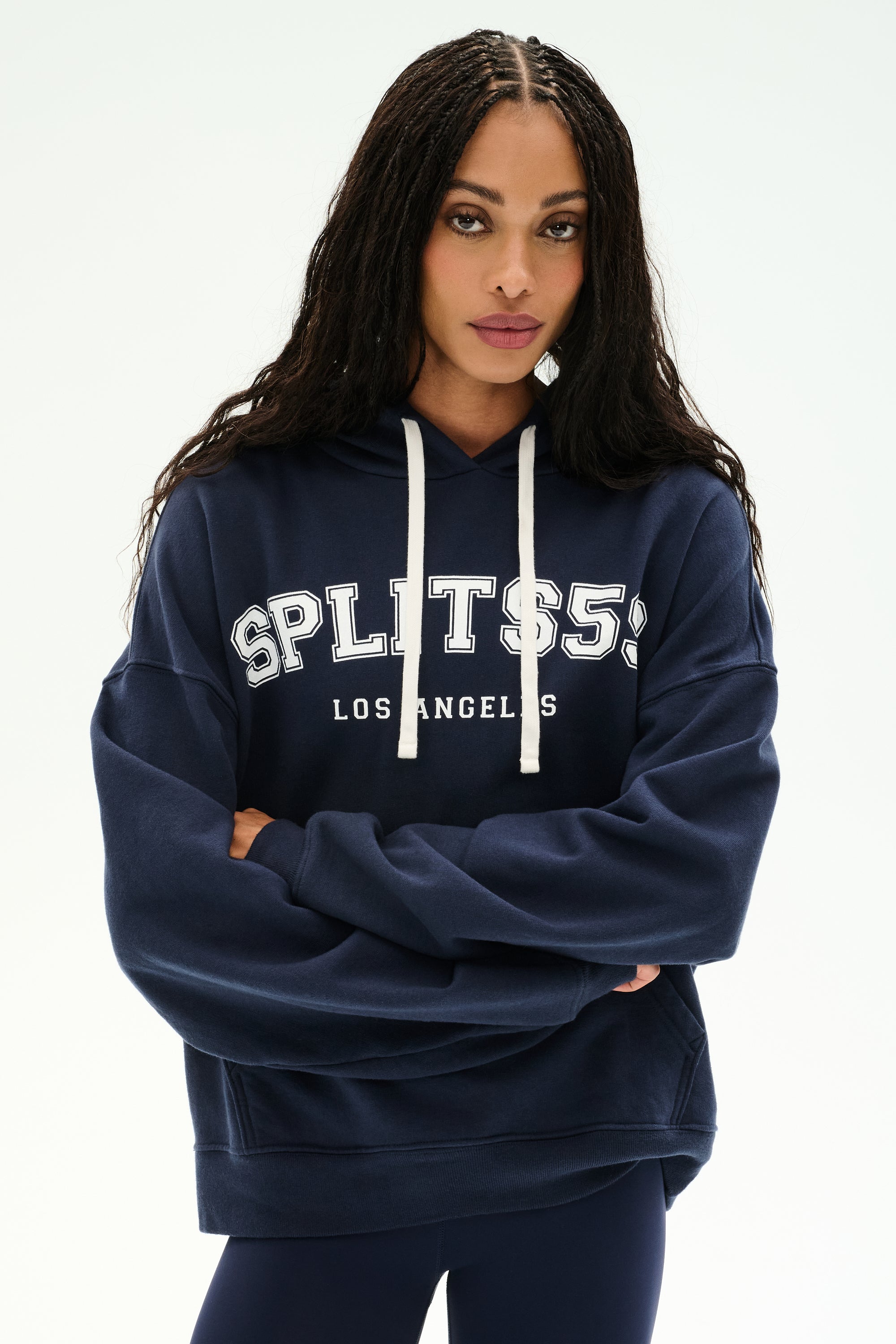 A woman stands with arms crossed against a plain background, wearing the SPLITS59 Stella French Terry Oversized Hoodie in Indigo—a soft 100% cotton piece featuring "SPLITS59 LOS ANGELES" printed on the front.