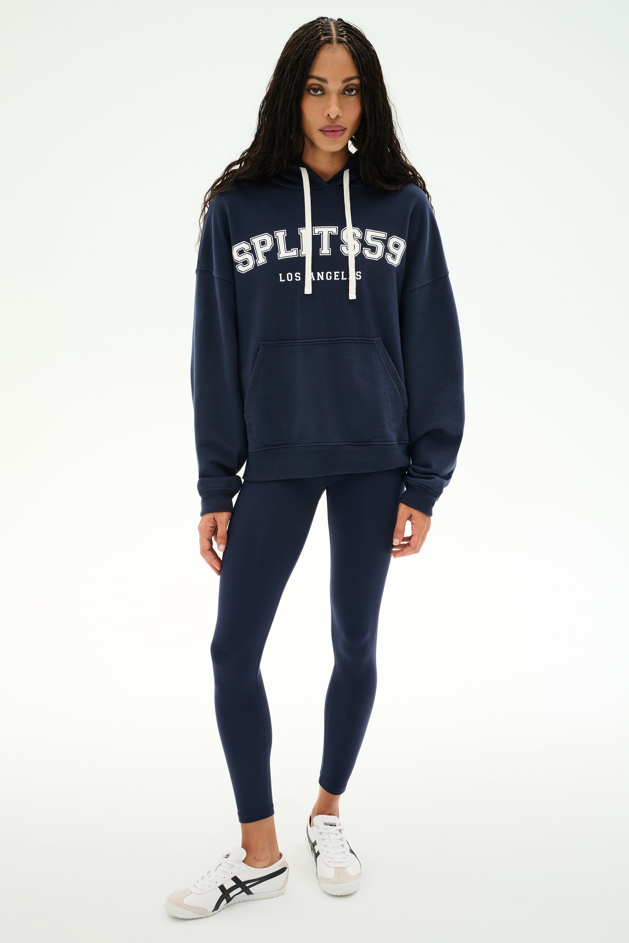 A person stands against a plain light background wearing the SPLITS59 Stella French Terry Oversized Hoodie in Indigo, featuring "SPLITS59 Los Angeles" branding, paired with navy leggings and white sneakers.
