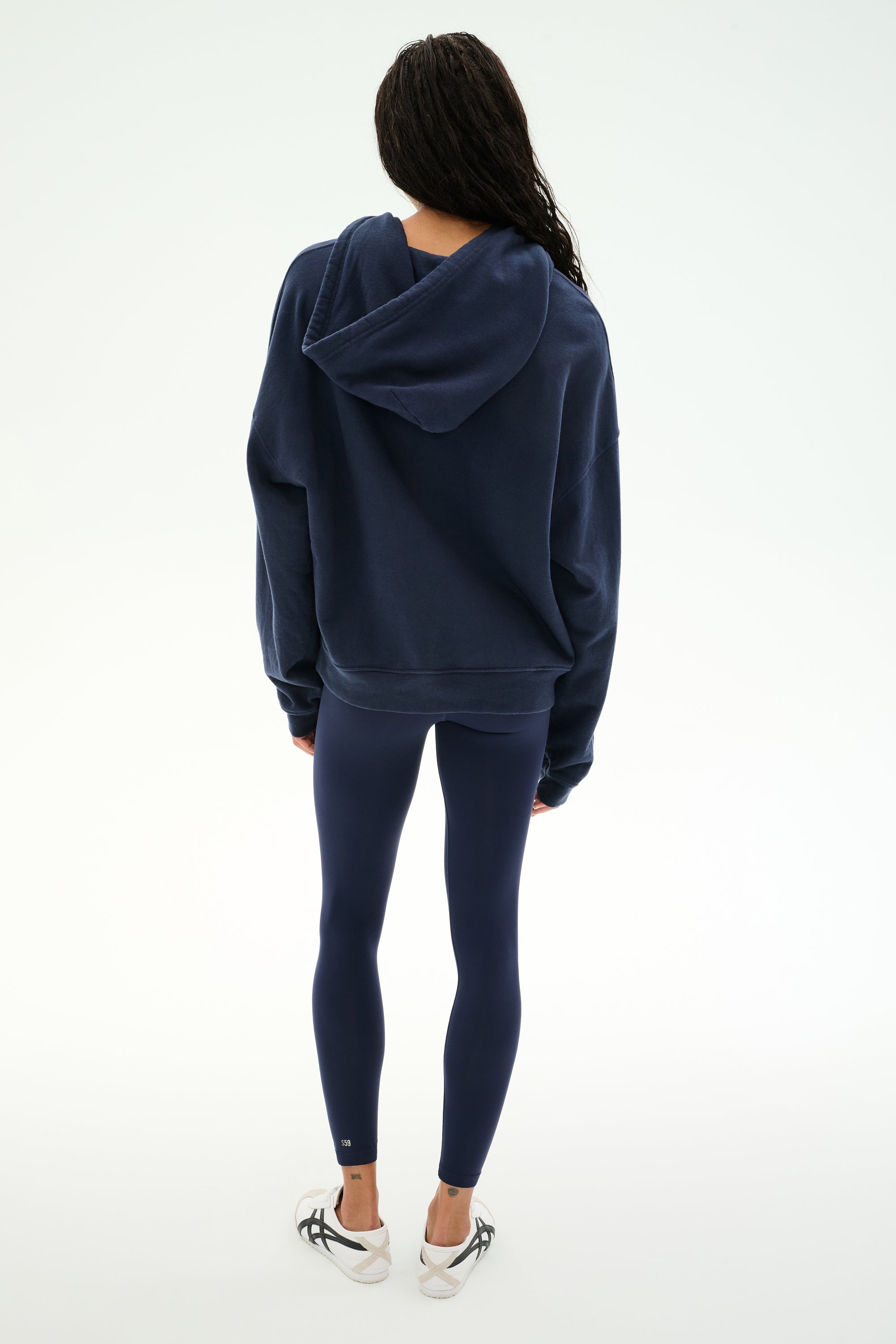 Standing with back to camera, the person is wearing the SPLITS59 Stella French Terry Oversized Hoodie in Indigo, paired with navy leggings and white sneakers against a plain white background.