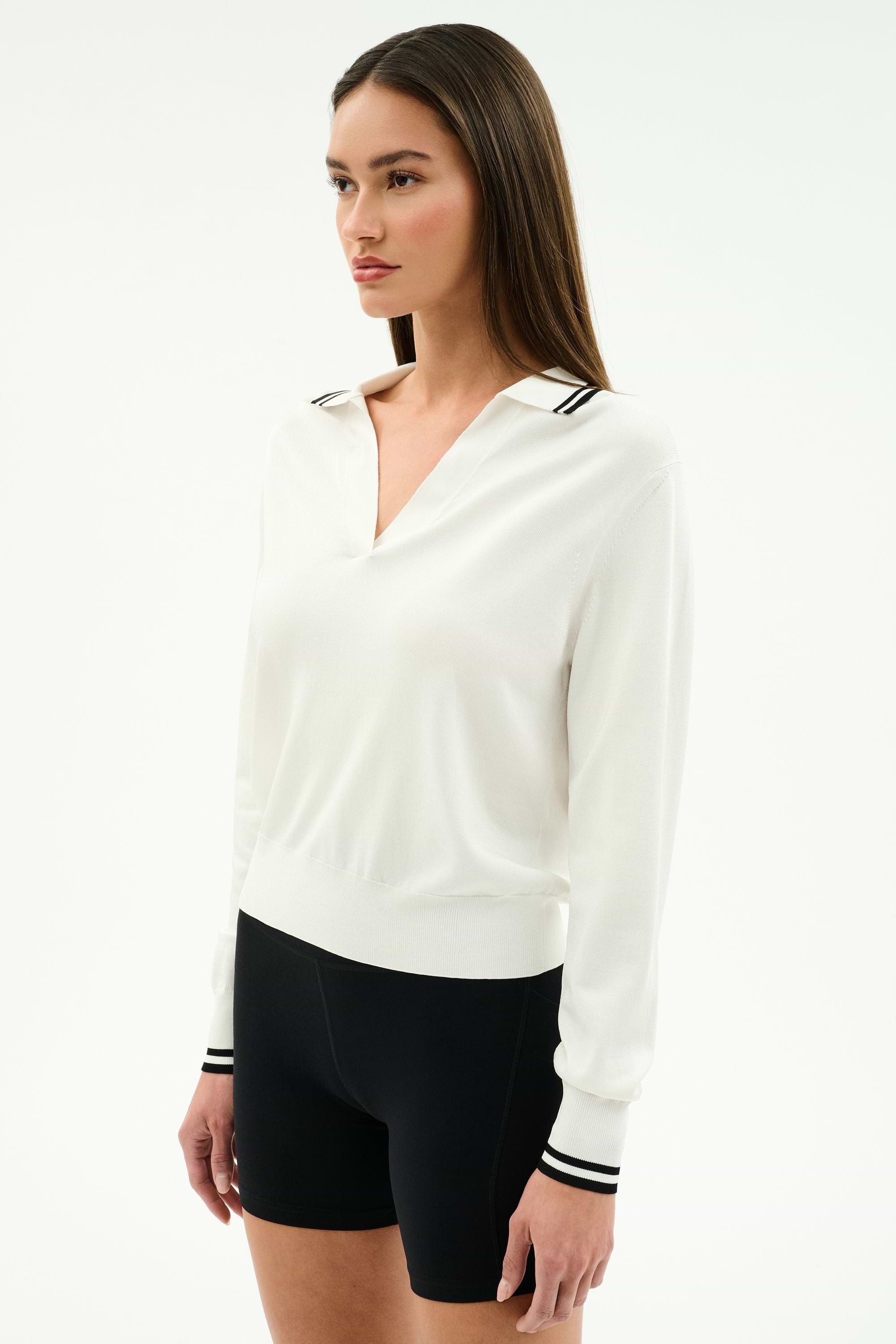 A woman poses against a plain background wearing the SPLITS59 Taylor Knit Polo in white with black stripes, featuring long sleeves and a collared design, styled with fitted black shorts.