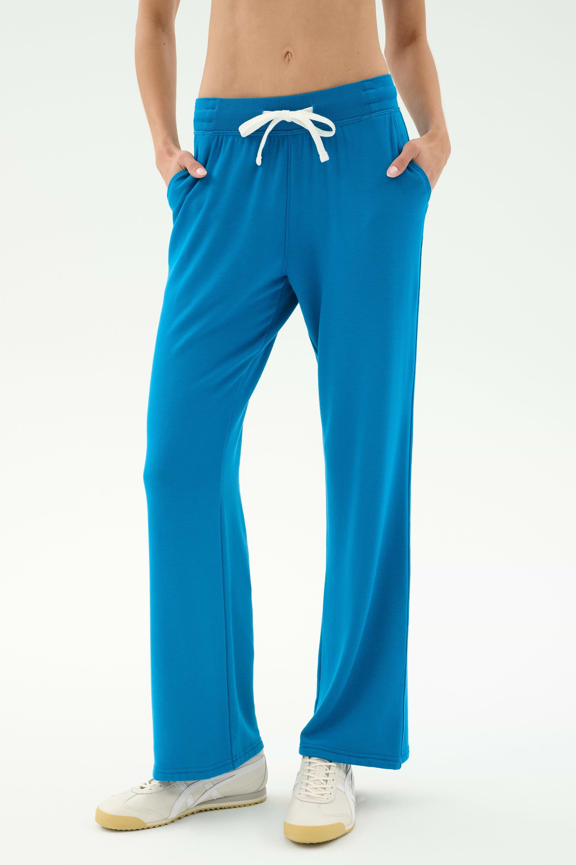 A person wears SPLITS59’s Raven Fleece Full Length Sweatpant in Stone Blue with white sneakers, standing hands-in-pockets against a plain light background.