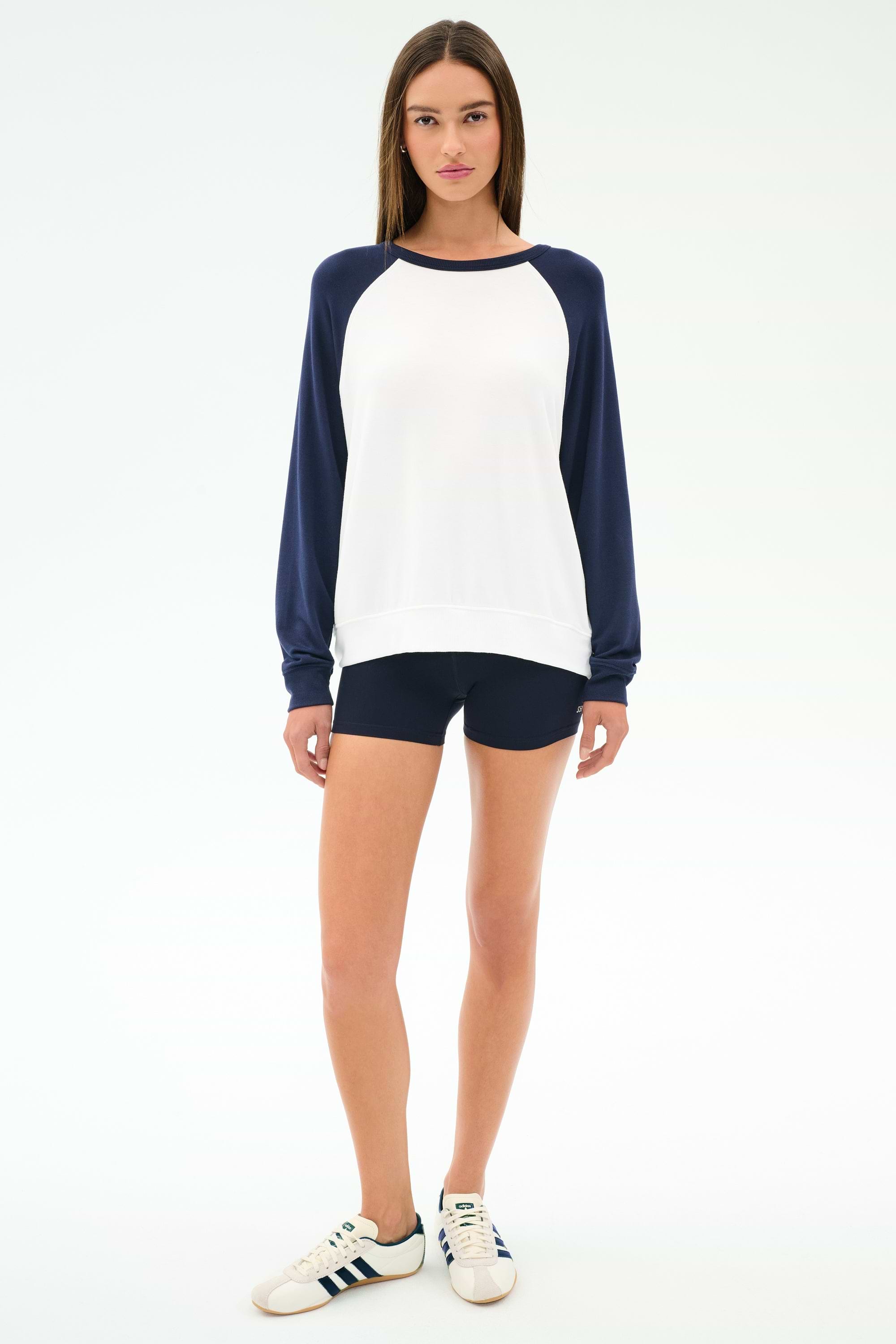 A woman faces forward on a plain white background wearing the SPLITS59 Andie Oversized Colorblock Fleece in Indigo/White, paired with navy modal spandex shorts and white Adidas sneakers.
