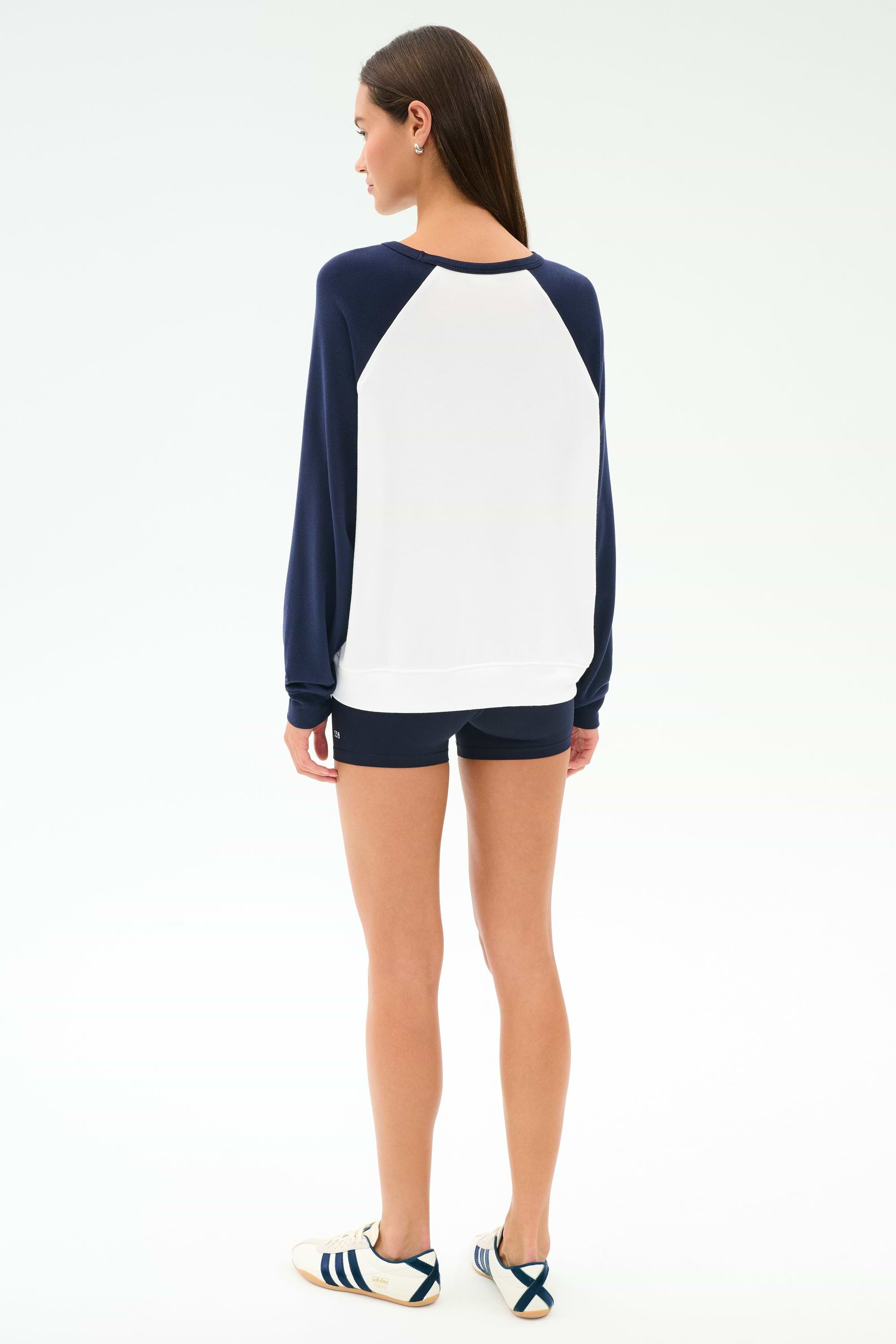 A person stands facing away, wearing the SPLITS59 Andie Oversized Colorblock Fleece in Indigo/White, black shorts, and white sneakers with navy stripes, against a plain white background.