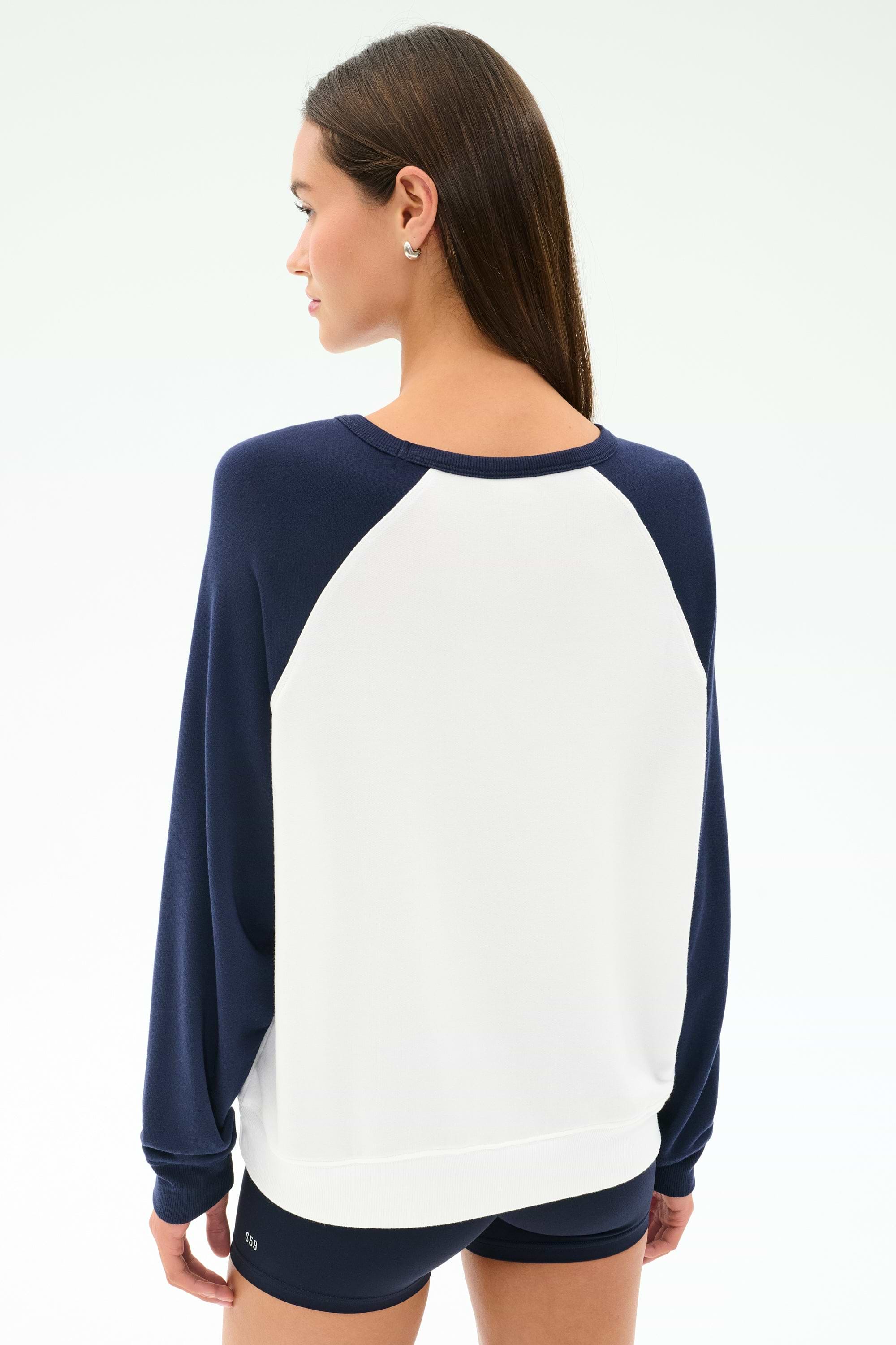 A woman with straight brown hair, seen from behind against a light background, wears the SPLITS59 Andie Oversized Colorblock Fleece in Indigo/White and black shorts.