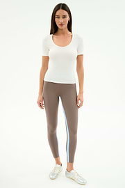 A woman faces forward wearing the SPLITS59 Ashby Rib Short Sleeve in white, taupe spandex-blend leggings with a blue stripe, and silver sneakers against a plain white background.