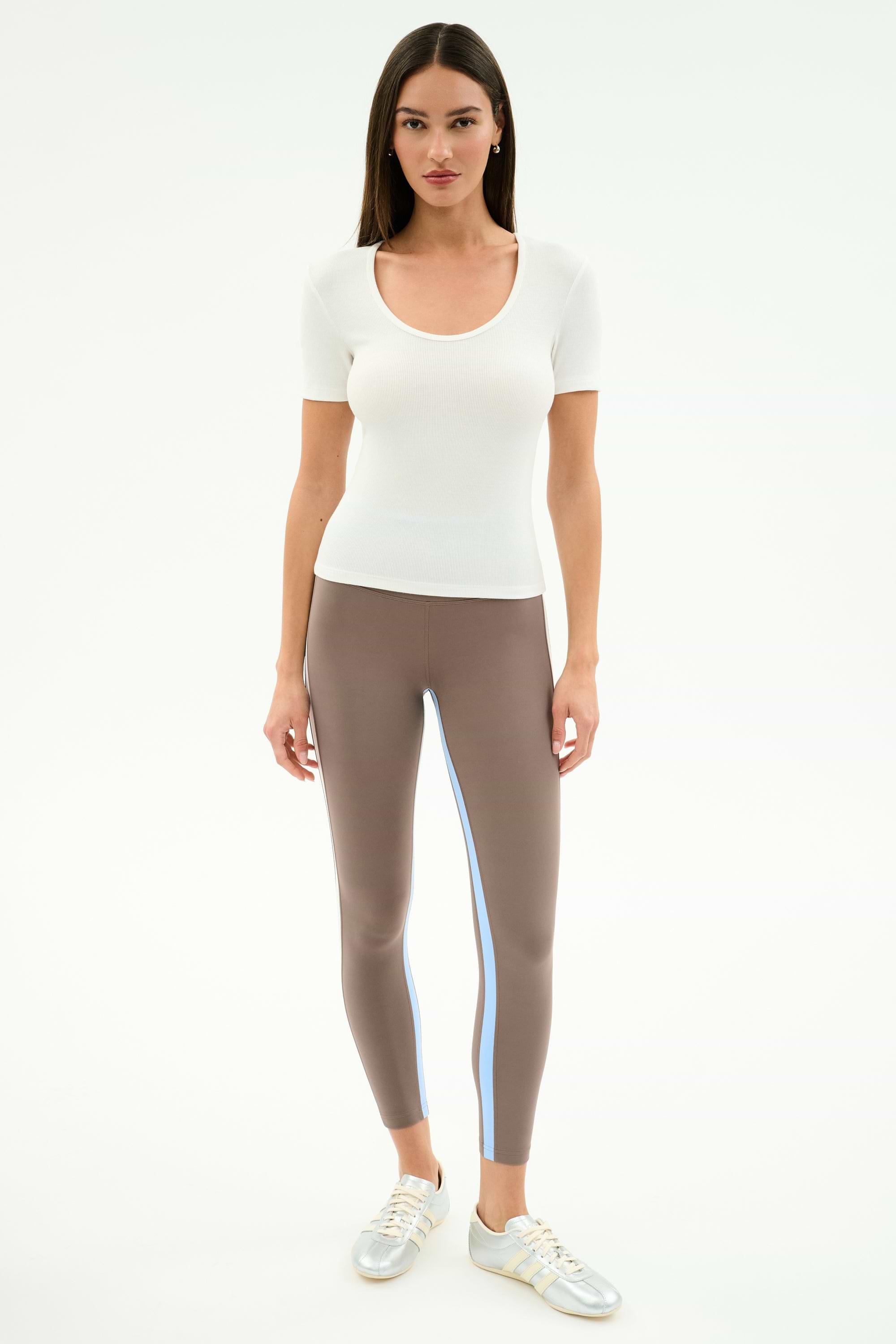 A woman faces forward wearing the SPLITS59 Ashby Rib Short Sleeve in white, taupe spandex-blend leggings with a blue stripe, and silver sneakers against a plain white background.