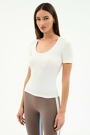 A woman with straight brown hair wears the SPLITS59 Ashby Rib Short Sleeve in white and taupe spandex blend leggings with a white side stripe, standing against a plain white background.