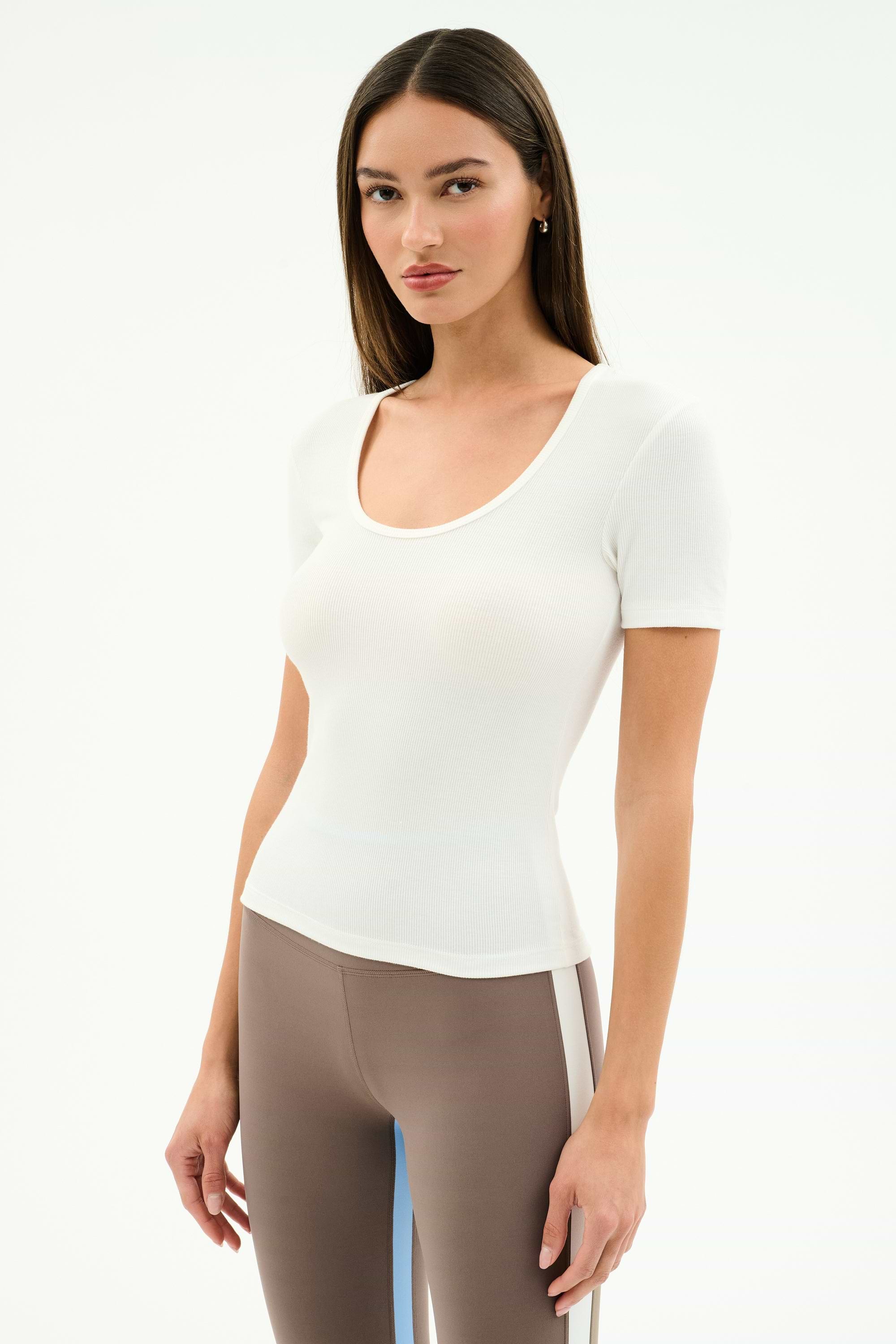 A woman with straight brown hair wears the SPLITS59 Ashby Rib Short Sleeve in white and taupe spandex blend leggings with a white side stripe, standing against a plain white background.
