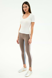 A woman faces forward wearing the SPLITS59 Ashby Rib Short Sleeve in white, taupe striped spandex-blend leggings, and silver sneakers against a plain light background.