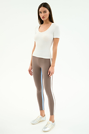 A woman faces forward wearing the SPLITS59 Ashby Rib Short Sleeve in white, taupe striped spandex-blend leggings, and silver sneakers against a plain light background.