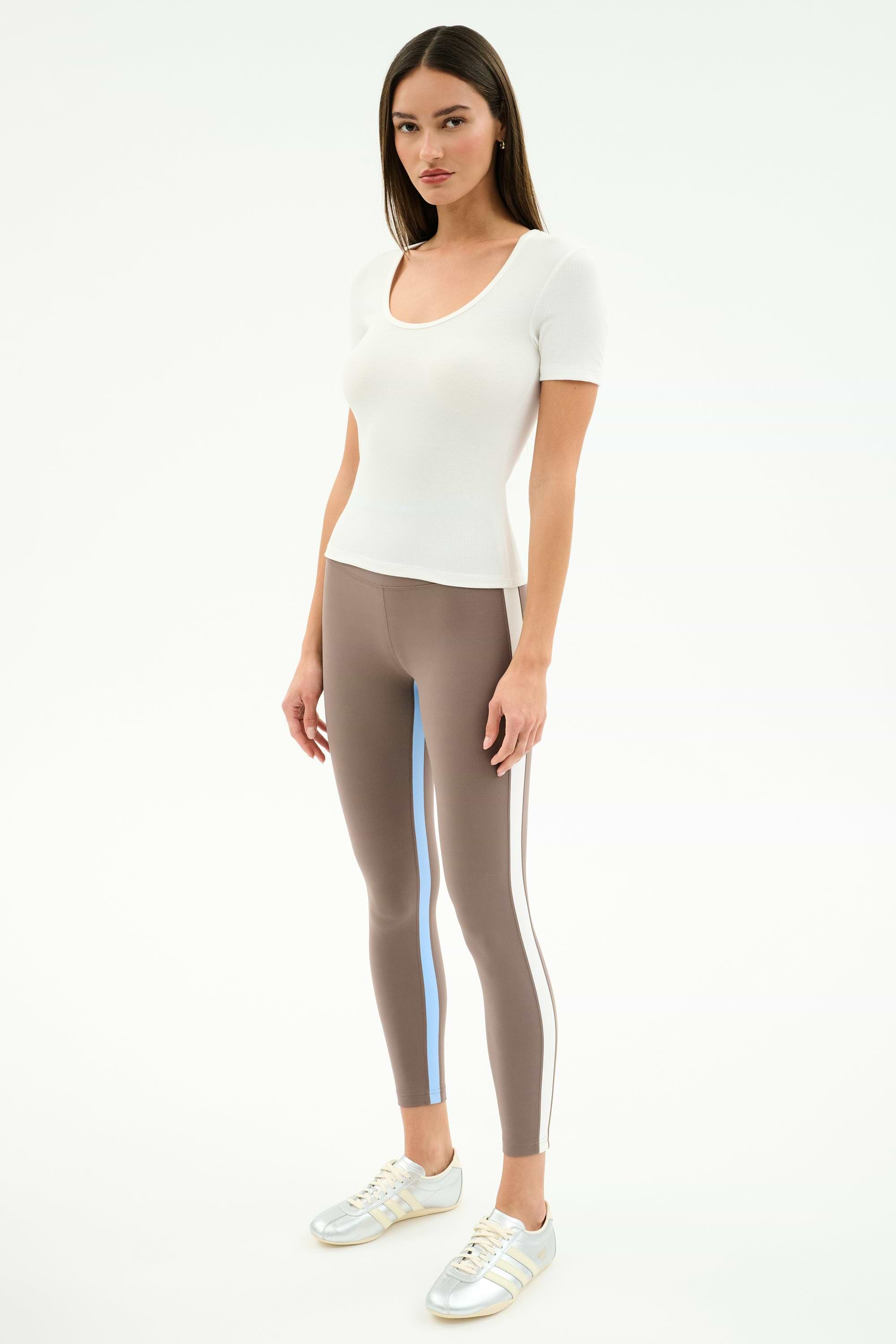A woman faces forward wearing the SPLITS59 Ashby Rib Short Sleeve in white, taupe striped spandex-blend leggings, and silver sneakers against a plain light background.