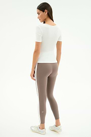 A woman stands against a white background, wearing the SPLITS59 Ashby Rib Short Sleeve in white and taupe leggings with a white side stripe, both in a comfy spandex blend, paired with white sneakers. She is turned slightly to the side.