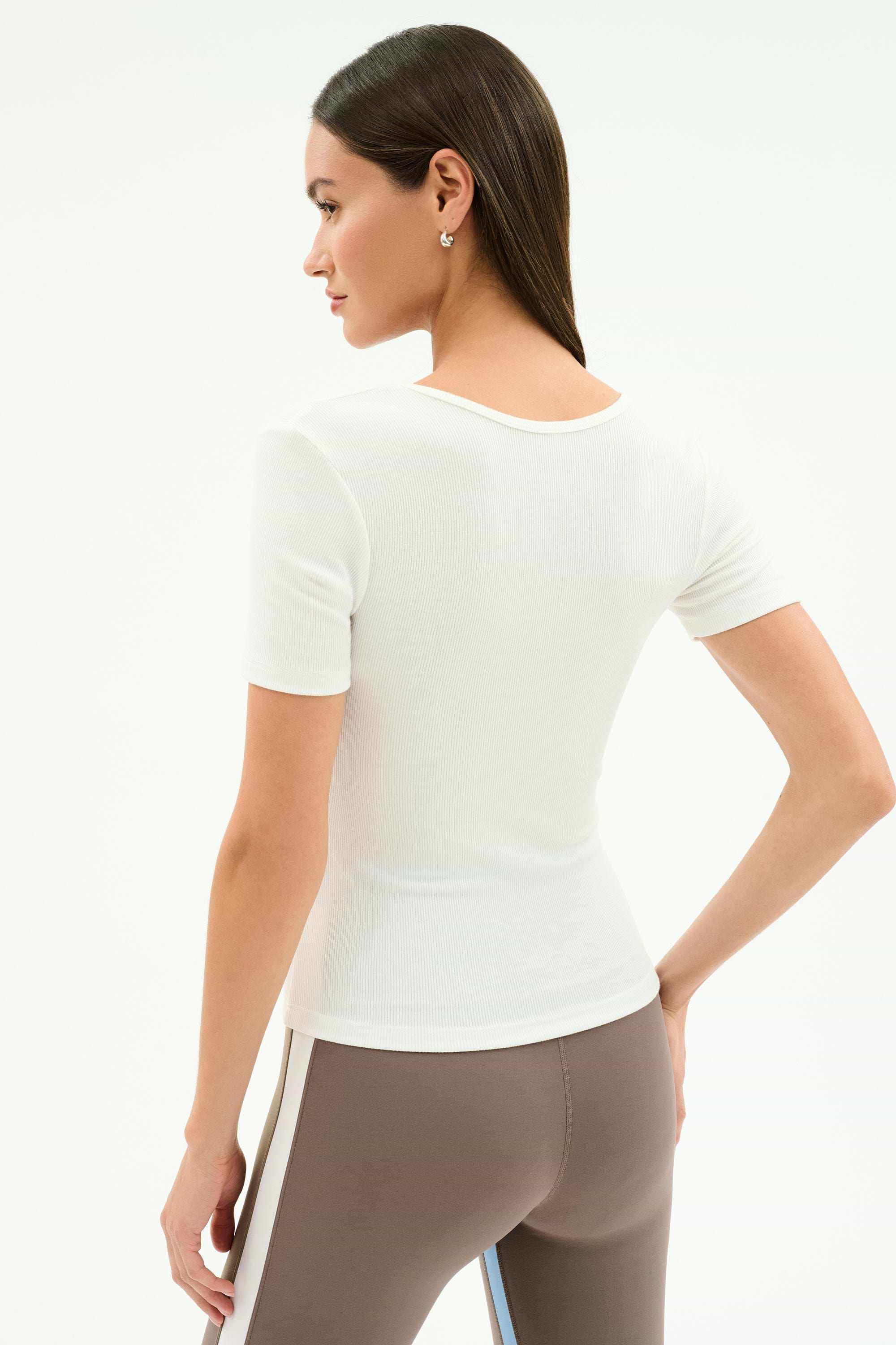 A woman stands with her back to the camera, wearing the SPLITS59 Ashby Rib Short Sleeve in white and brown spandex blend leggings with side stripes, set against a plain background.