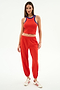 A woman stands against a white background wearing SPLITS59’s Kiki Rib Crop Tank in Lotus Red/Cobalt, red jogger pants, and white sneakers with red stripes.