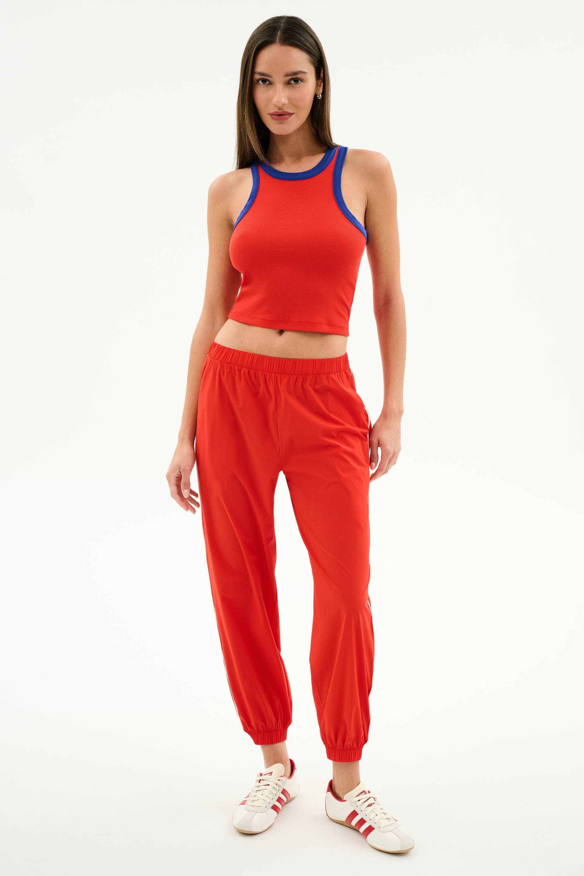A woman stands against a white background wearing SPLITS59’s Kiki Rib Crop Tank in Lotus Red/Cobalt, red jogger pants, and white sneakers with red stripes.