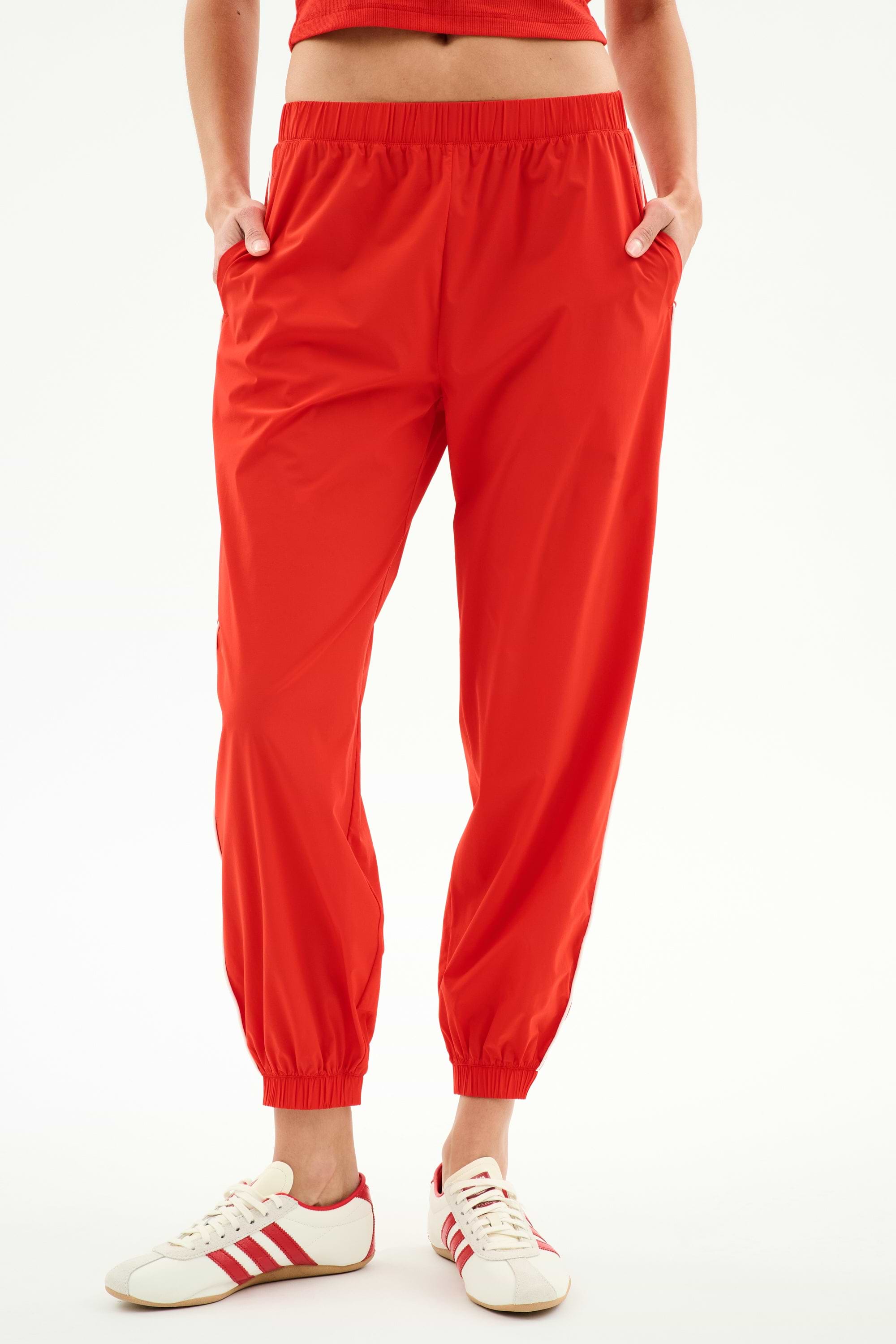 Model wears the SPLITS59 Megan Woven Nylon Jogger w/ Piping in Lotus Red/Creme, paired with white sneakers featuring red stripes, standing hands-in-pockets against a plain background.
