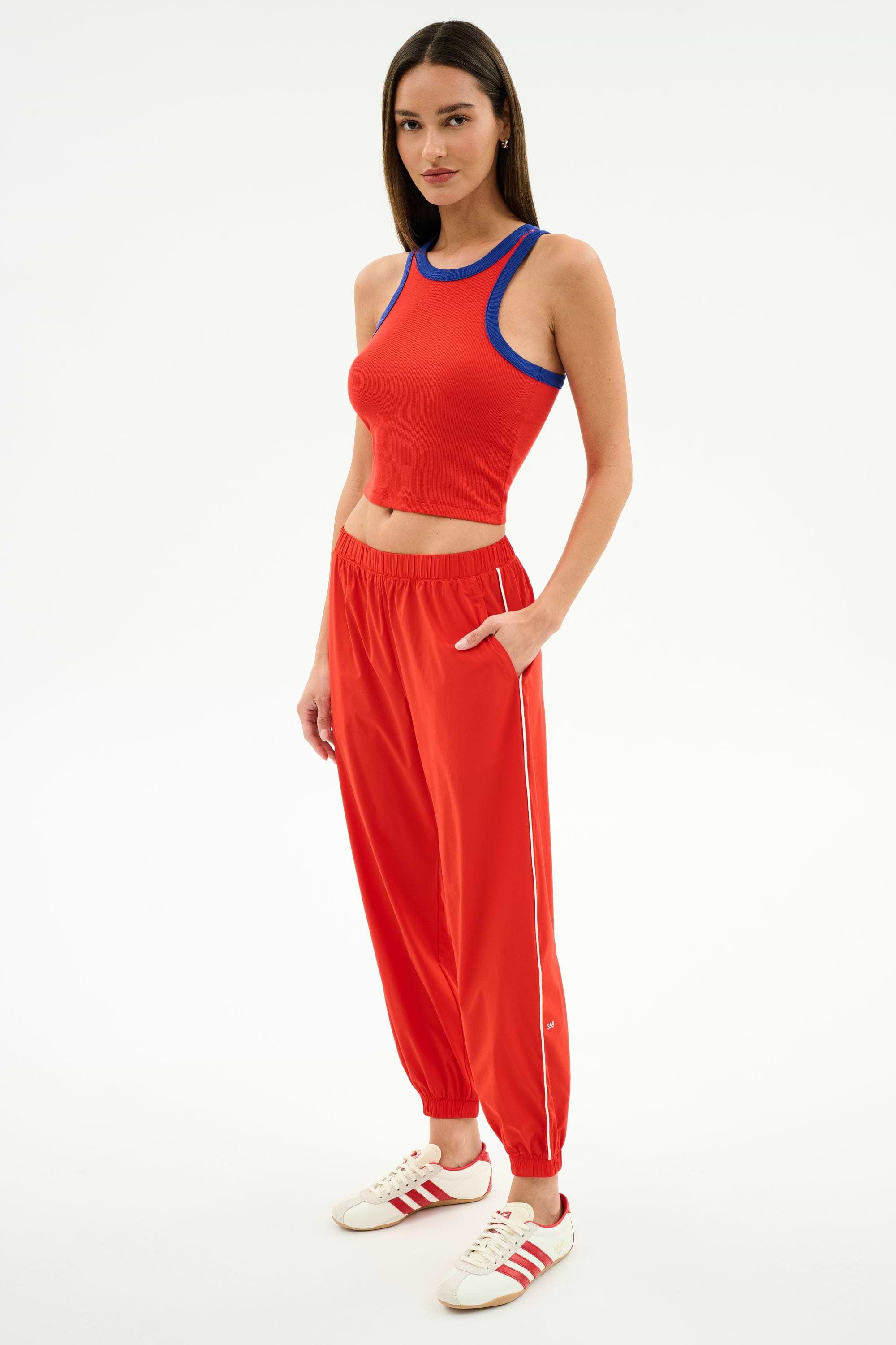 A woman wears the SPLITS59 Kiki Rib Crop Tank in Lotus Red/Cobalt, paired with red track pants with a white stripe and white sneakers with red accents—an ideal outfit for gym sessions.