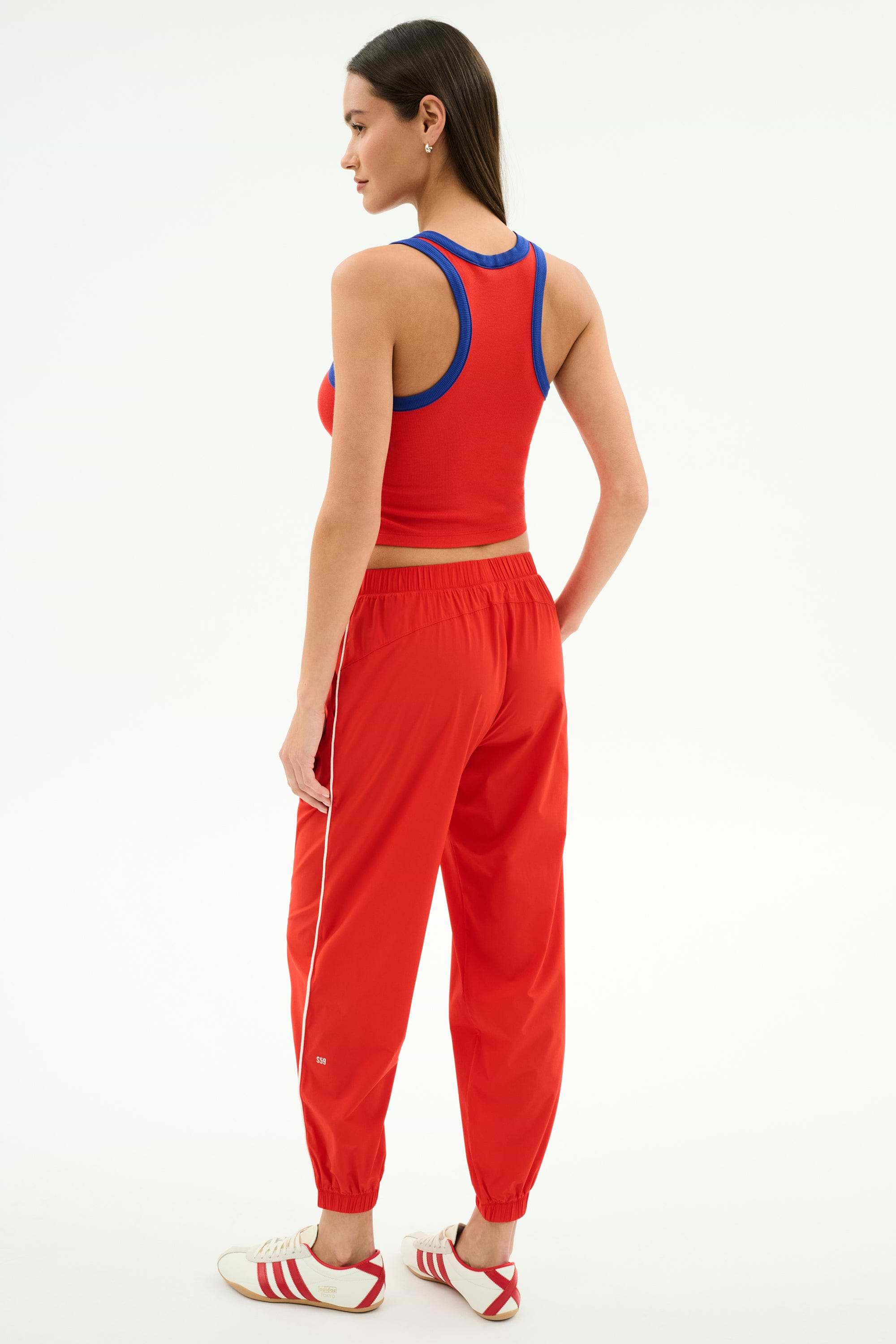 A woman faces away, wearing the SPLITS59 Kiki Rib Crop Tank in Lotus Red/Cobalt—perfect for gym workouts—with red track pants featuring white stripes and white Adidas sneakers with red accents.
