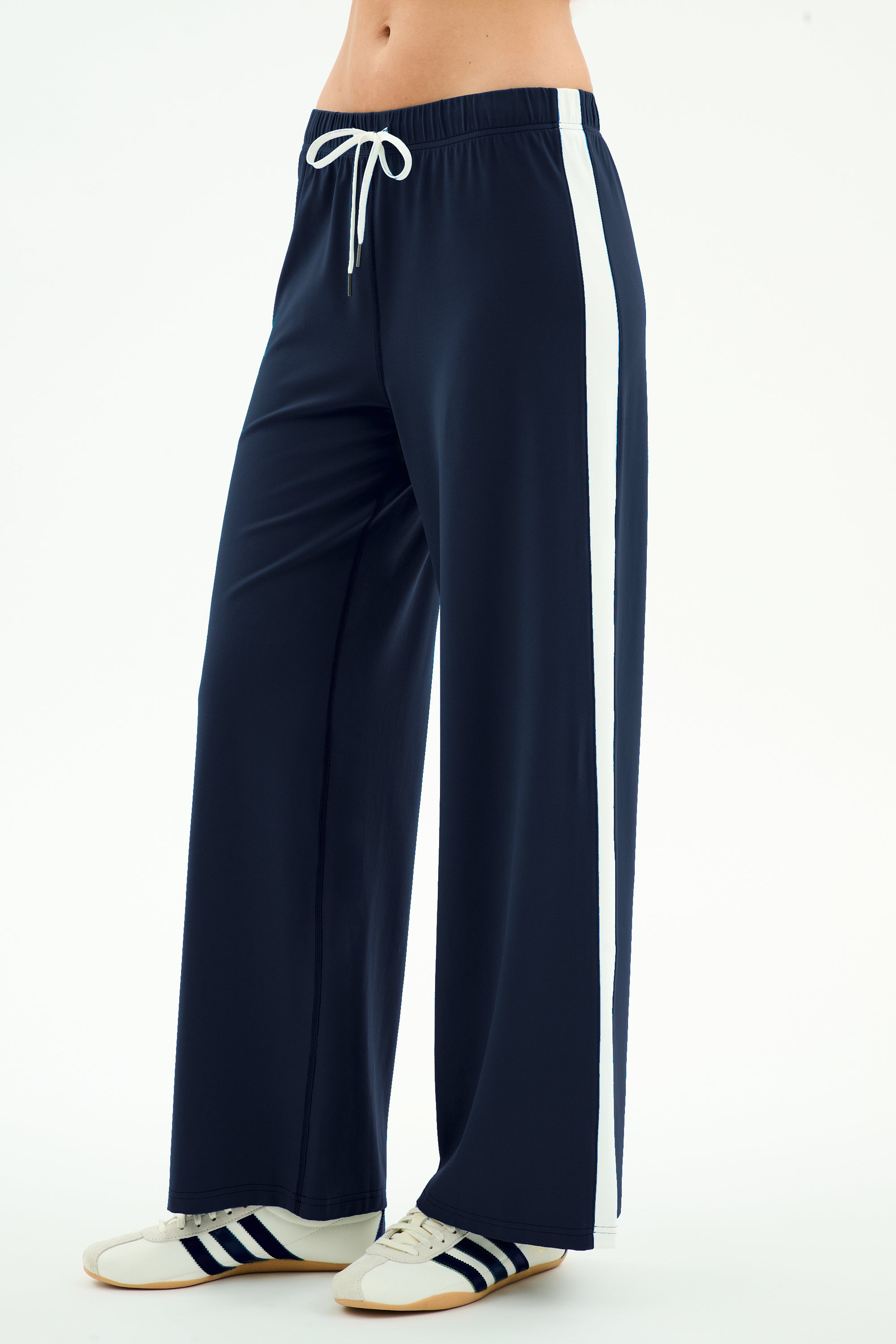 A person stands against a plain light background wearing the SPLITS59 Willow Airweight Wide Leg pants in Indigo/White—navy blue pants with a white side stripe made from stretch activewear fabric—and white sneakers with navy stripes.