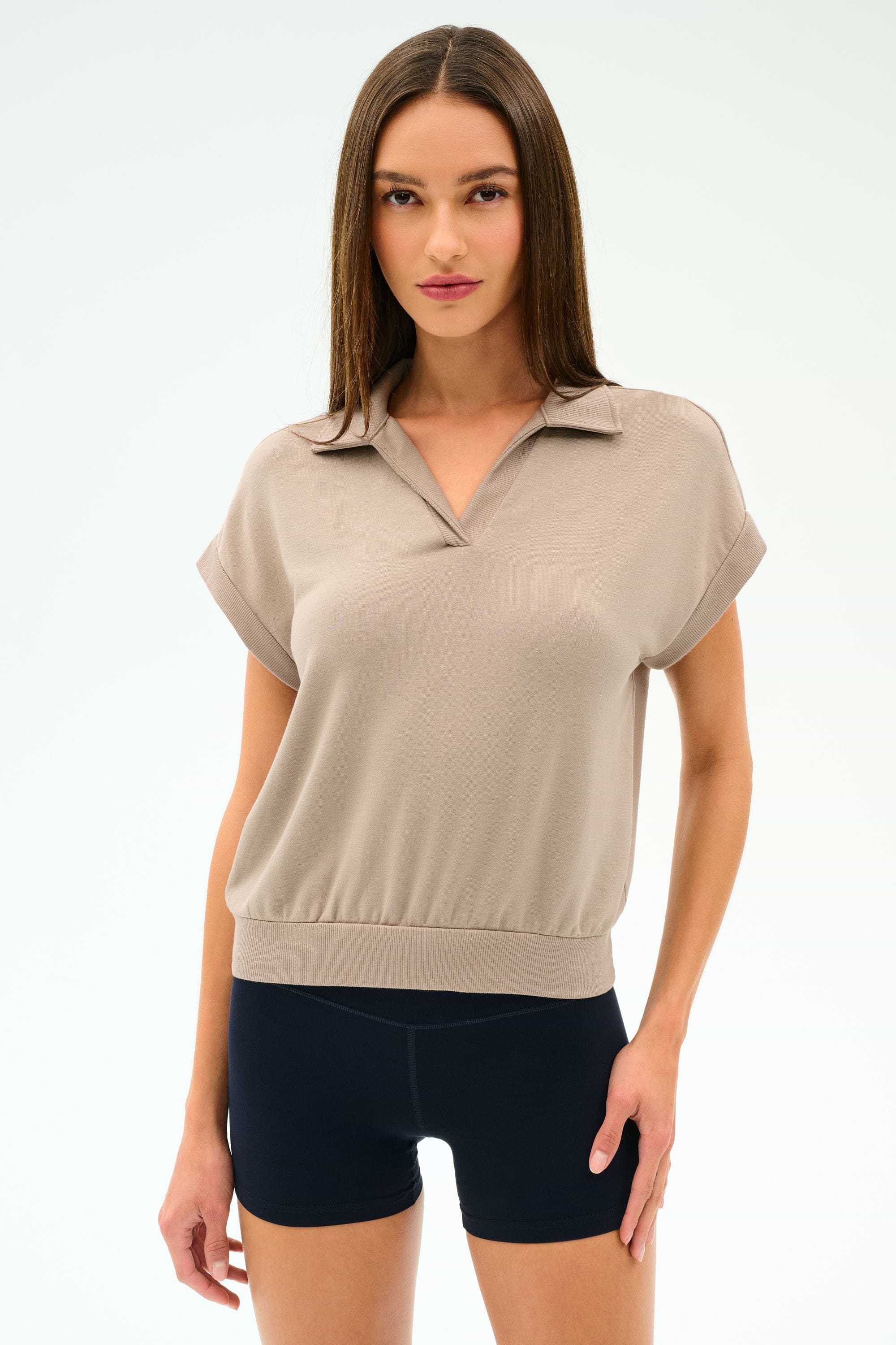A woman poses against a plain background wearing the SPLITS59 Evie Fleece Sleeveless Polo in Latte and black fitted stretch shorts.