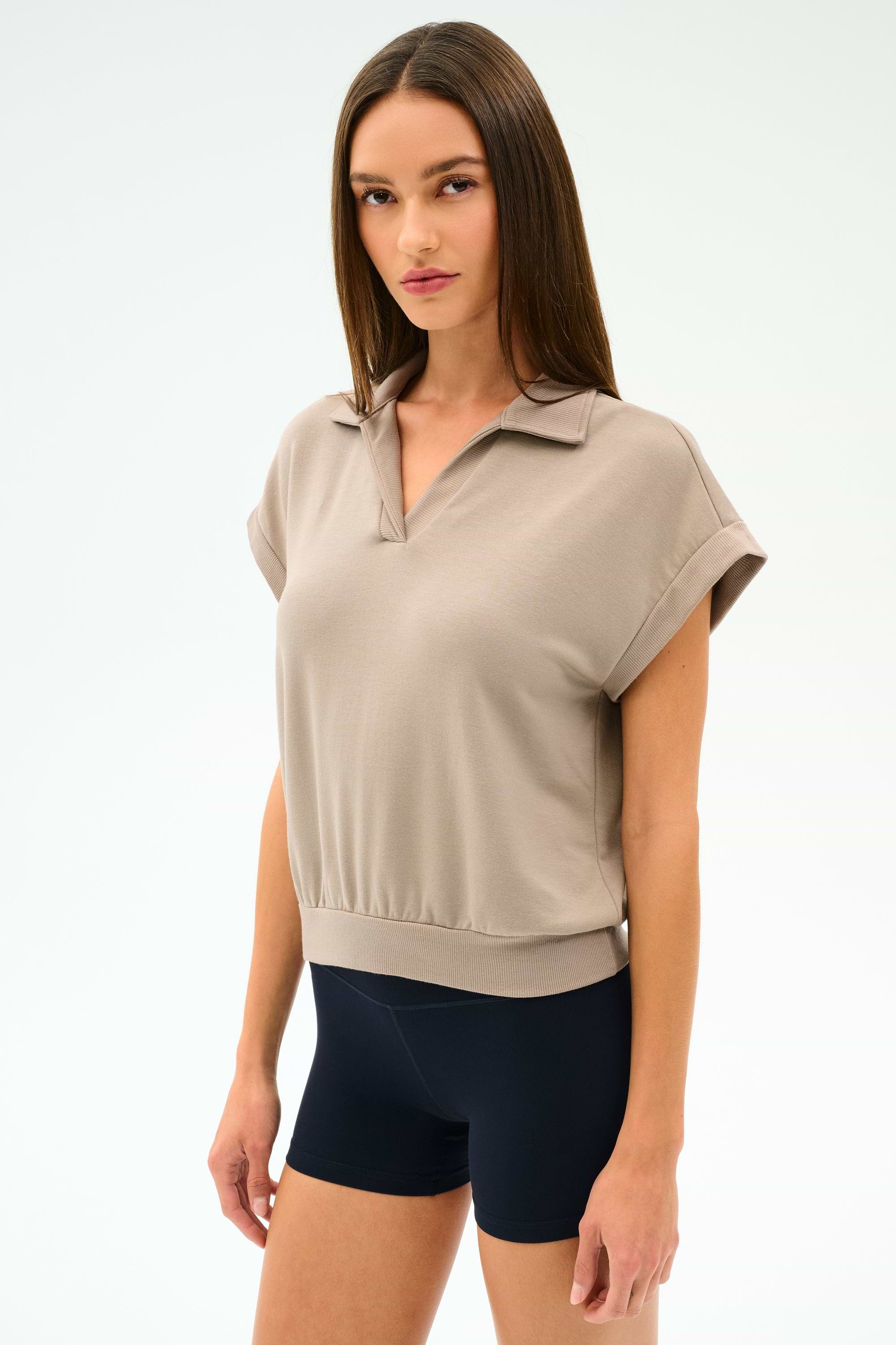 A woman with straight brown hair wears the SPLITS59 Evie Fleece Sleeveless Polo in Latte and black fitted stretch shorts, standing against a plain light background.