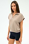 A woman with straight brown hair wears the SPLITS59 Evie Fleece Sleeveless Polo in Latte and black fitted stretch shorts, standing against a plain light background.