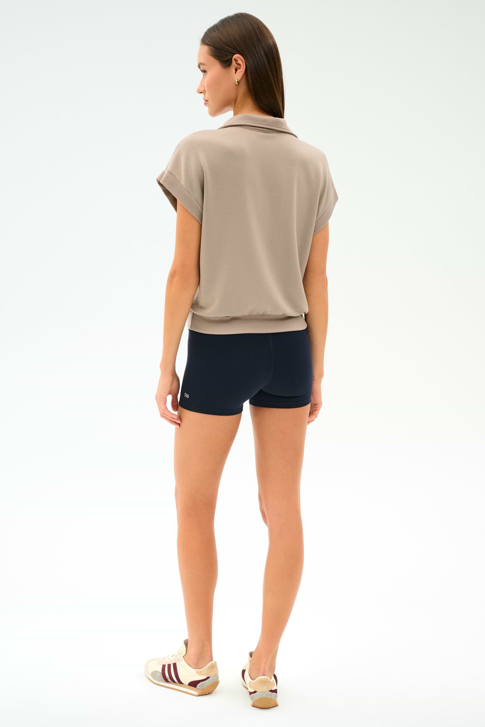 A woman stands facing away, wearing the SPLITS59 Evie Fleece Sleeveless Polo in Latte, black shorts, and beige sneakers with burgundy stripes against a plain white background.
