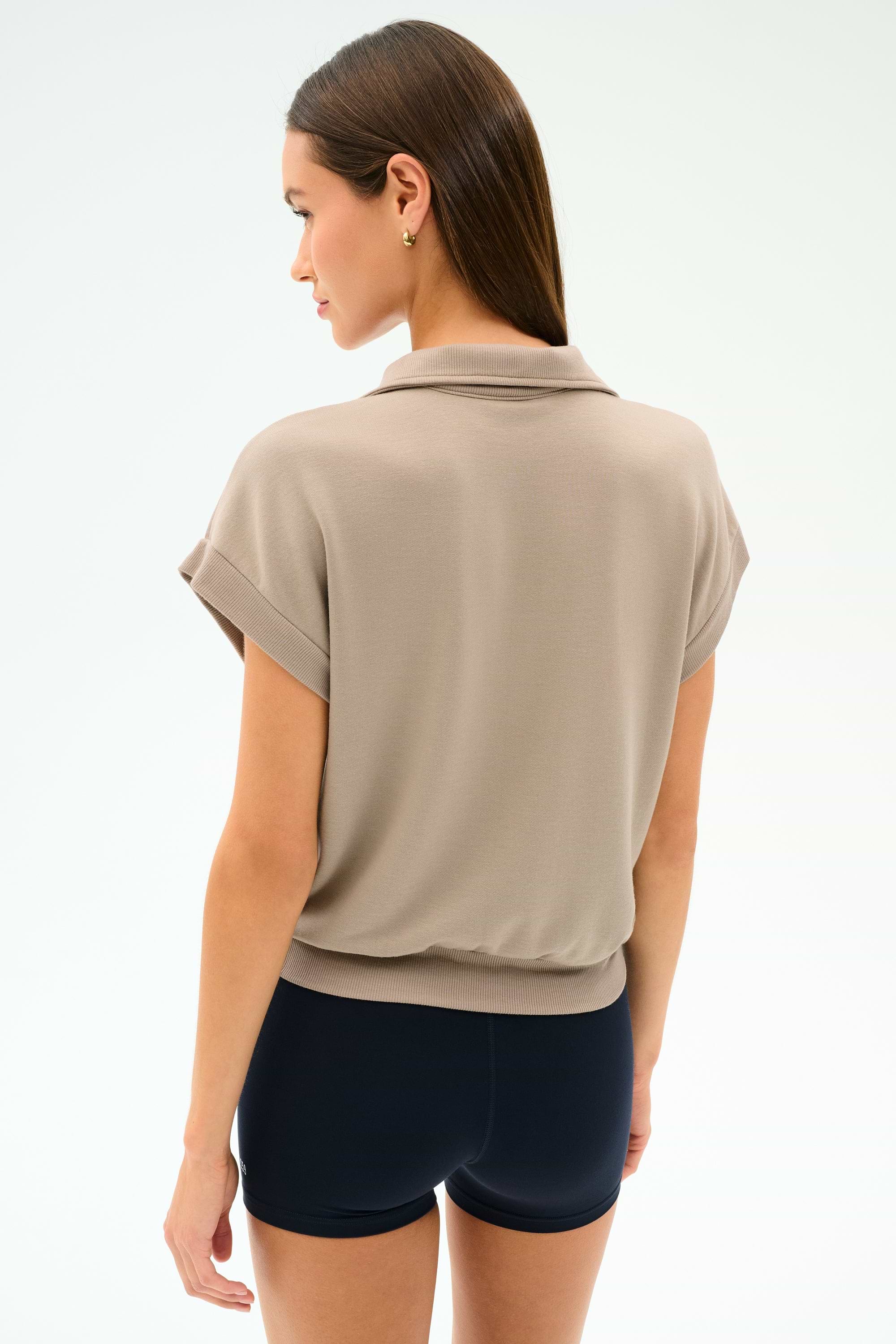 A woman stands with her back turned, wearing the SPLITS59 Evie Fleece Sleeveless Polo in Latte against a plain light background.