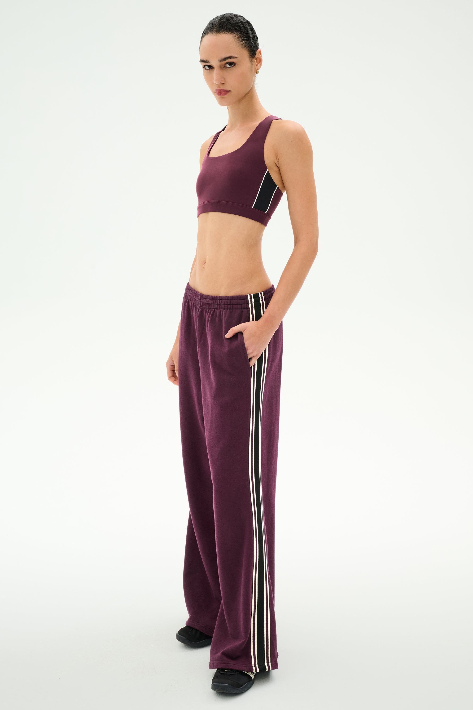 A person stands against a plain white background wearing SPLITS59′s Dallas French Terry Wide Leg Sweatpant in Dark Cherry/Black, paired with a matching maroon 100% cotton sports bra featuring white side stripes.
