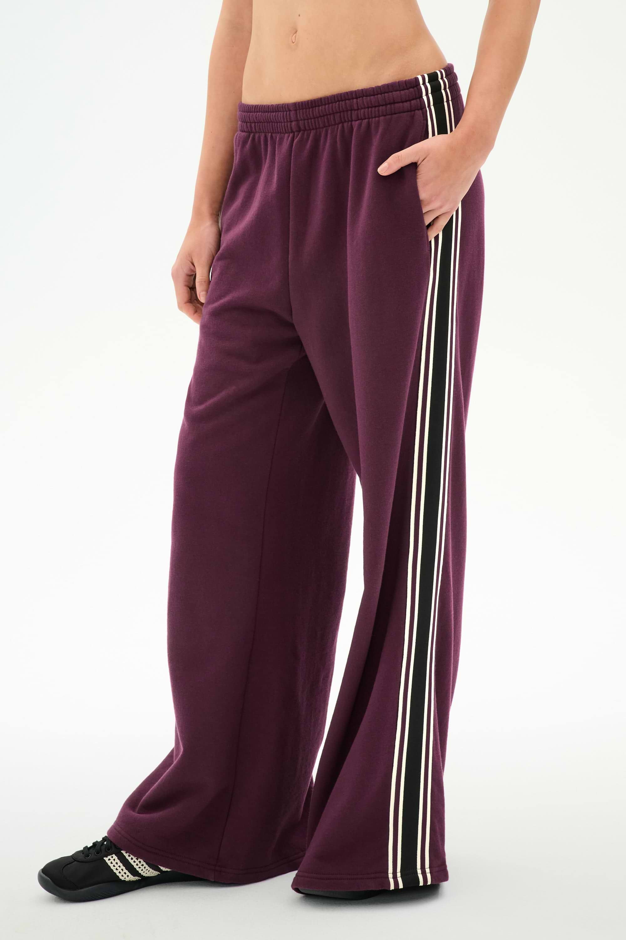 Wearing SPLITS59 Dallas French Terry Wide Leg Sweatpant in Dark Cherry/Black—a loose-fitting, 100% cotton design with white and black side stripes—this person also has a matching top partially visible and black shoes, standing against a plain white background.