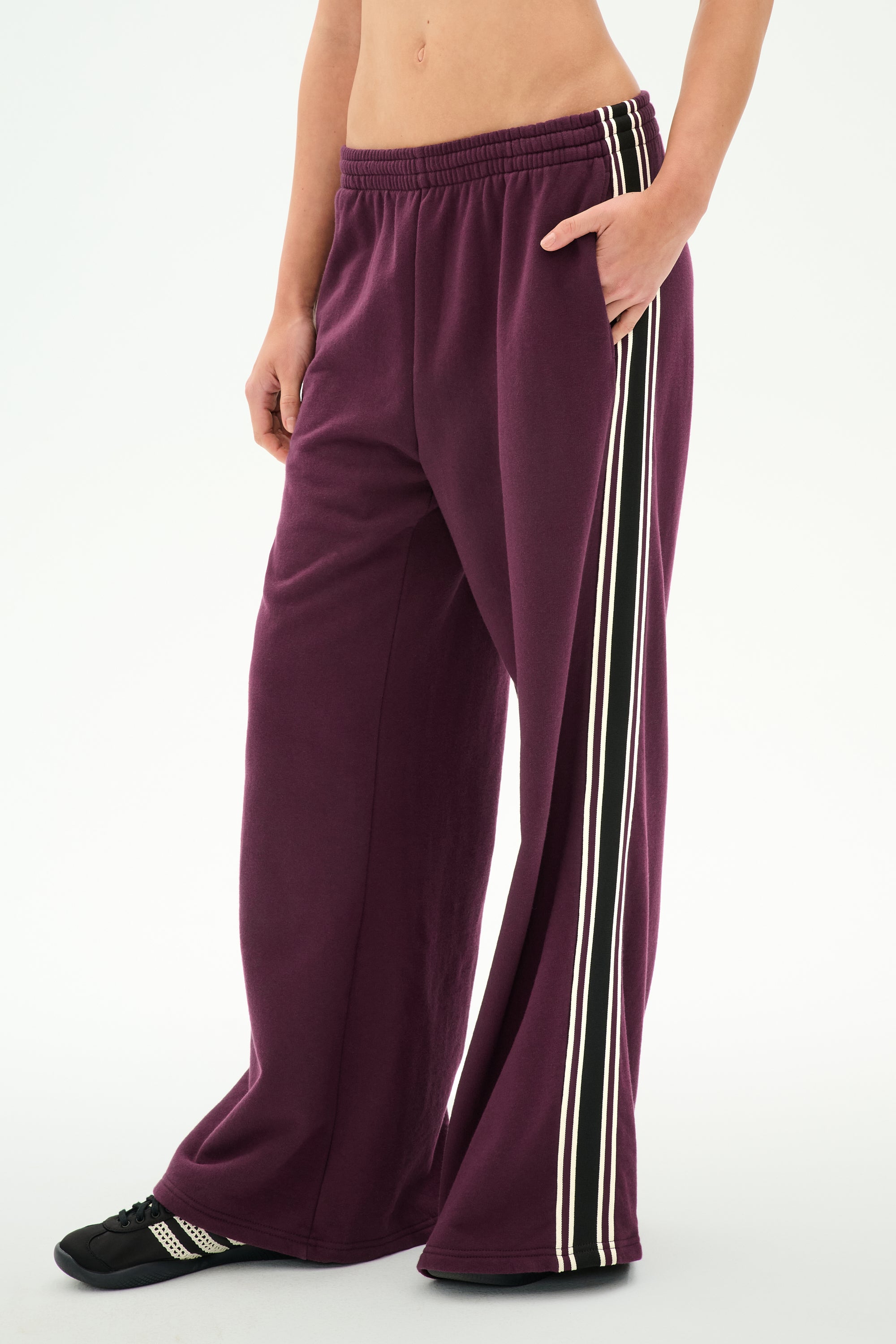 Wearing SPLITS59 Dallas French Terry Wide Leg Sweatpant in Dark Cherry/Black—a loose-fitting, 100% cotton design with white and black side stripes—this person also has a matching top partially visible and black shoes, standing against a plain white background.