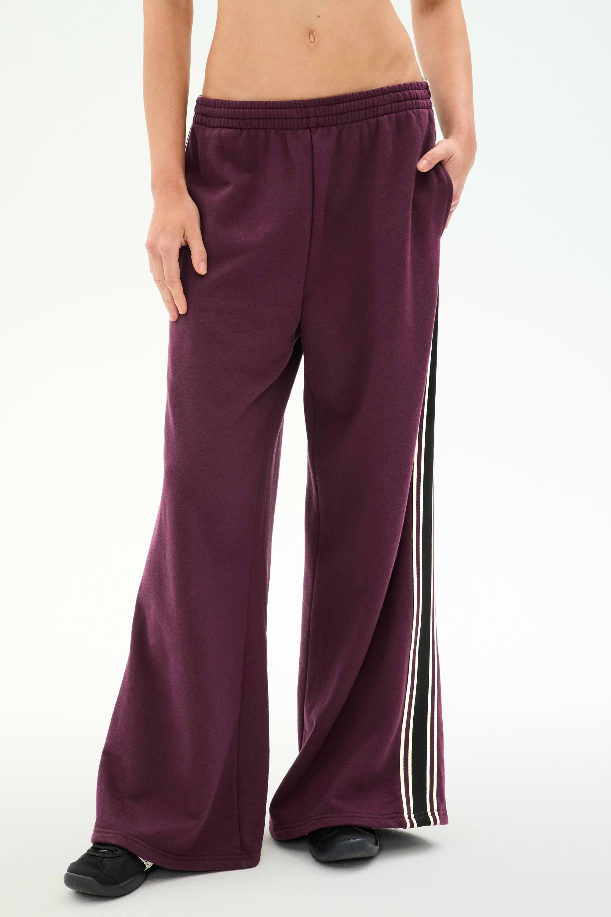 A person wearing SPLITS59’s Dallas French Terry Wide Leg Sweatpant in Dark Cherry/Black—loose-fitting 100% cotton sweatpants featuring black and white side stripes—stands with one hand in a pocket.