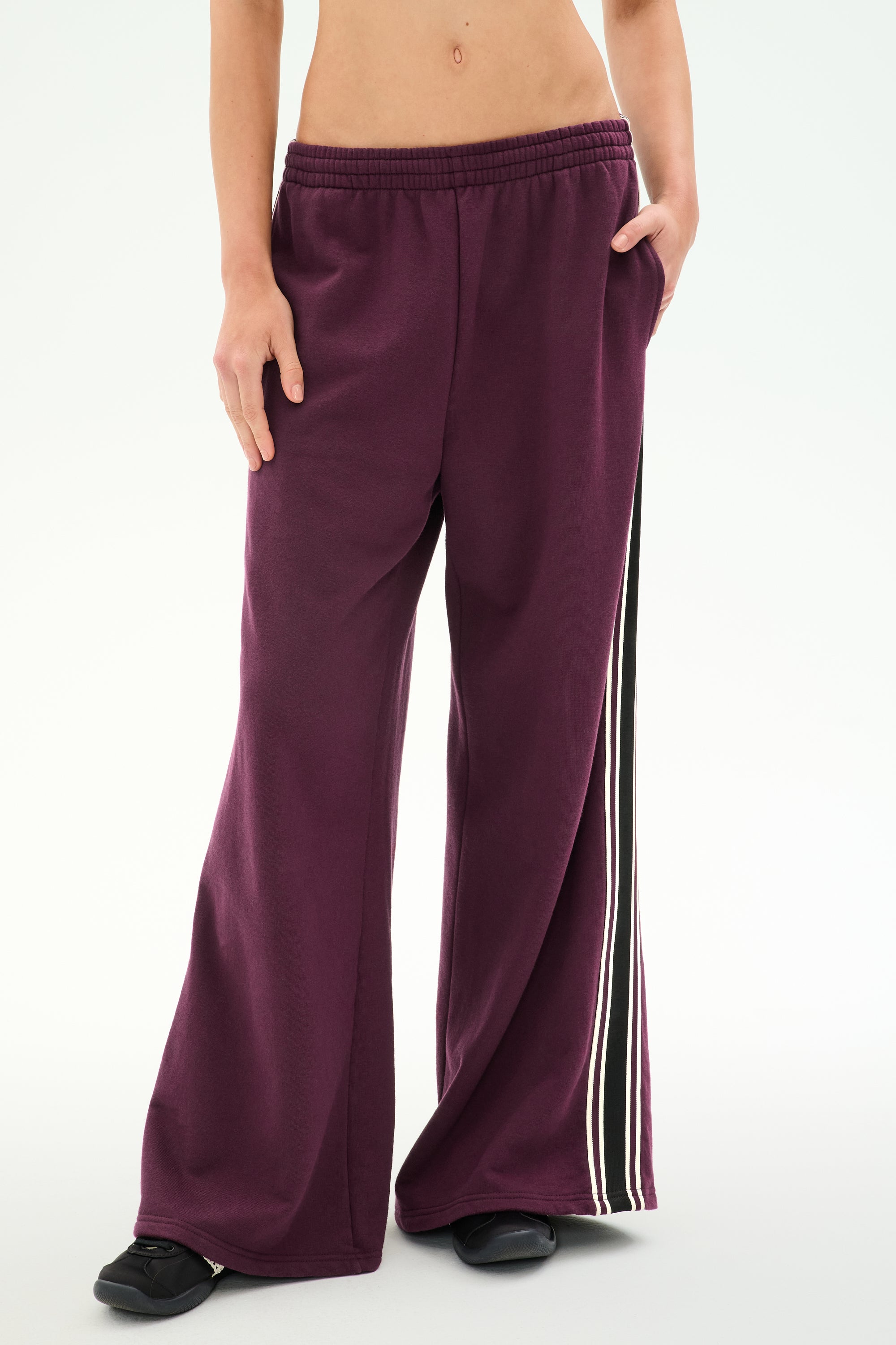 A person wearing SPLITS59’s Dallas French Terry Wide Leg Sweatpant in Dark Cherry/Black—loose-fitting 100% cotton sweatpants featuring black and white side stripes—stands with one hand in a pocket.