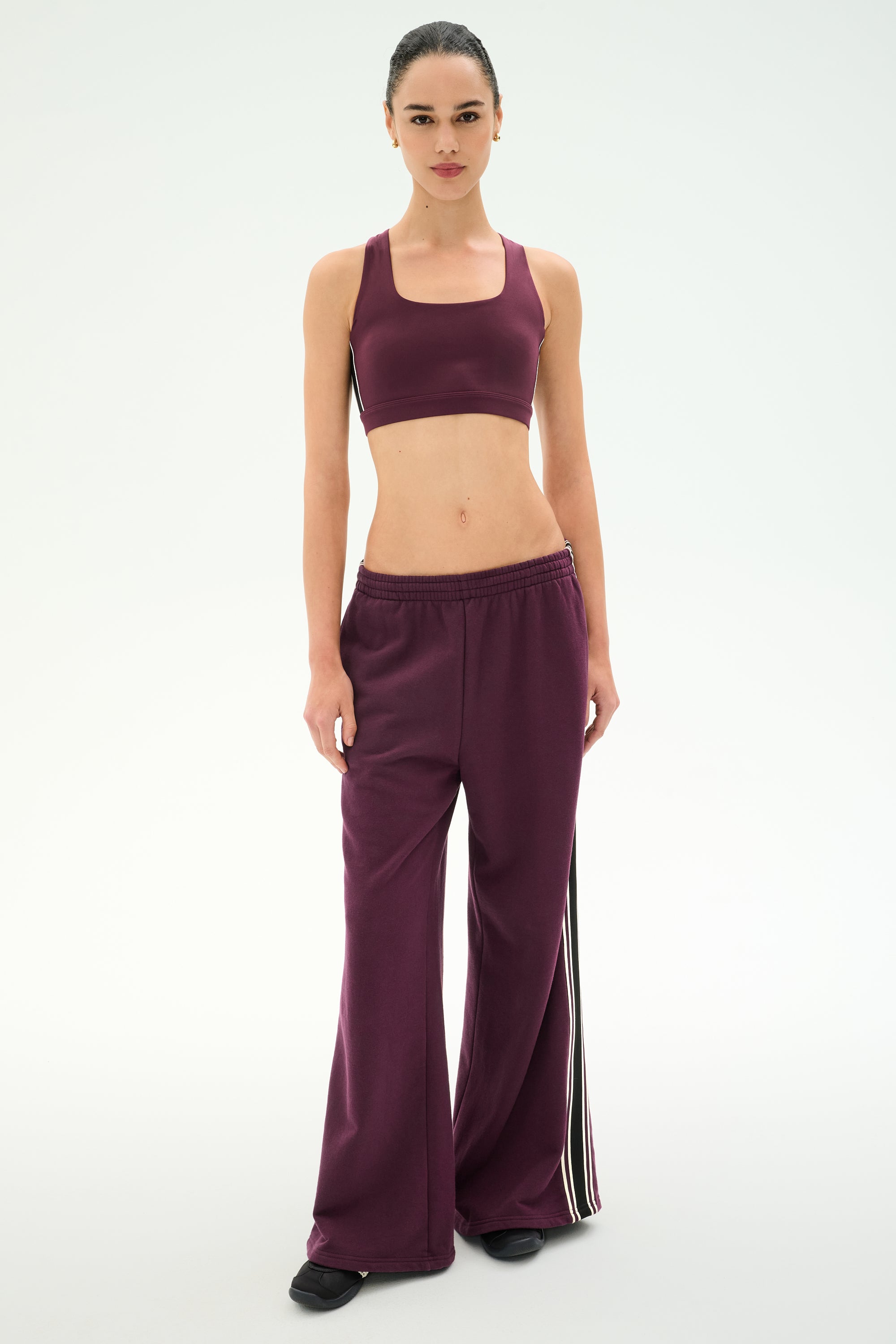 A person stands facing forward wearing SPLITS59’s Dallas French Terry Wide Leg Sweatpant in Dark Cherry/Black, featuring matching wide-leg track pants with white side stripes, crafted from soft 100% cotton, against a plain light background.