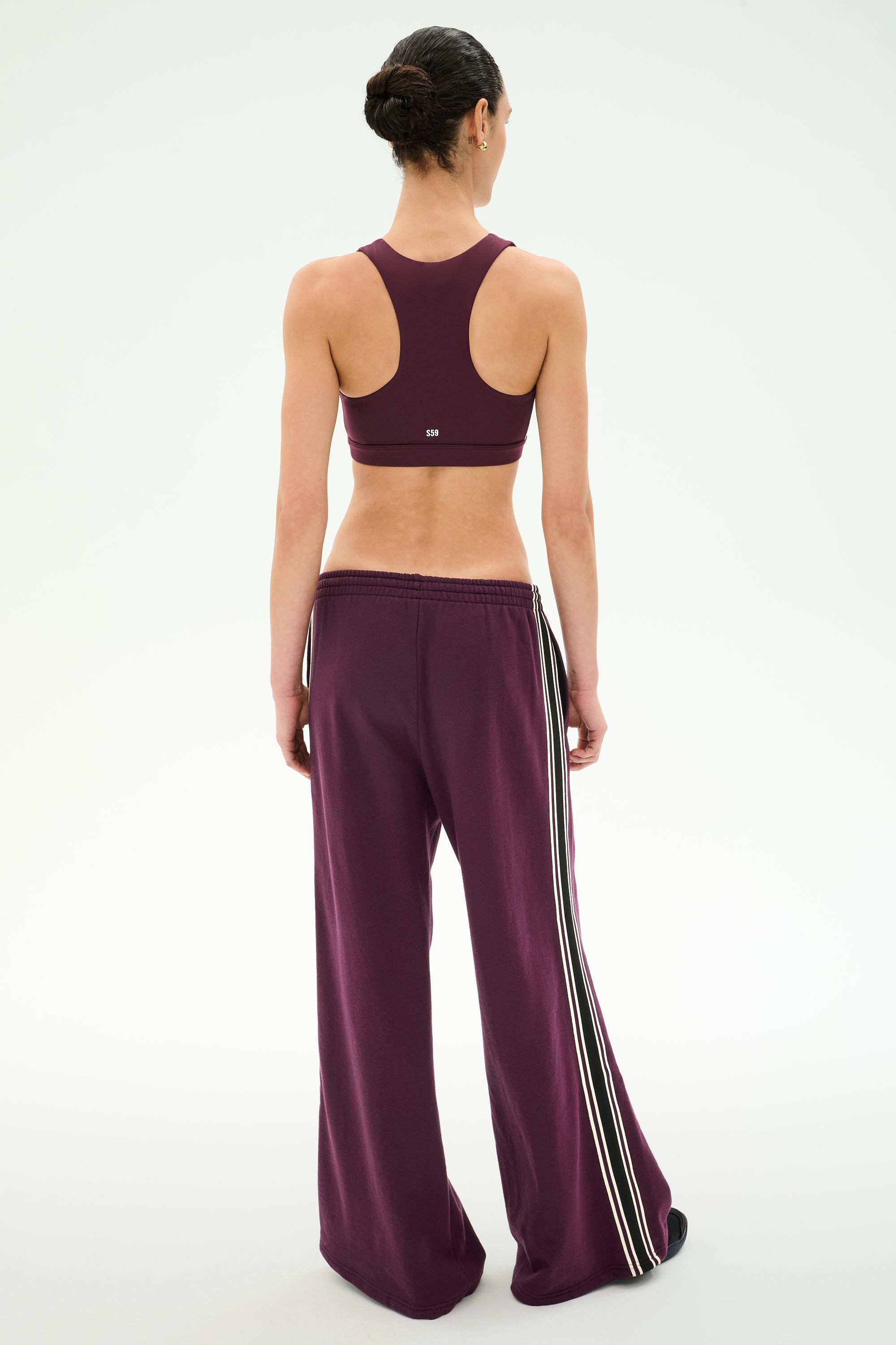 A person stands with their back to the camera, wearing SPLITS59’s Dallas French Terry Wide Leg Sweatpant in Dark Cherry/Black—crafted from 100% cotton and featuring side stripes—paired with a matching maroon sports bra, set against a plain white background.