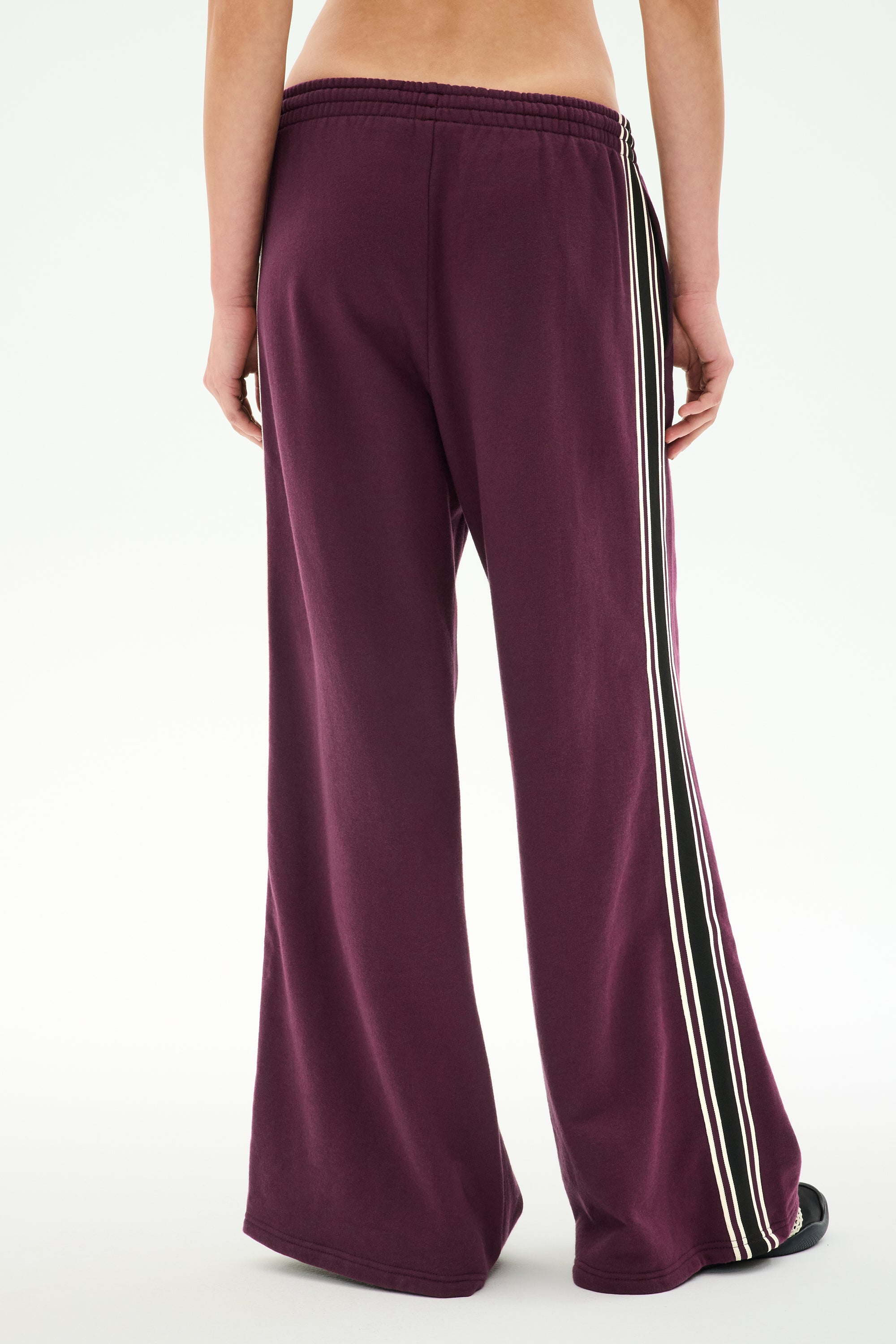 A person is shown from the back, standing against a plain background and wearing the SPLITS59 Dallas French Terry Wide Leg Sweatpant in Dark Cherry/Black—a loose-fitting pair of 100% cotton maroon pants featuring black and white side stripes.