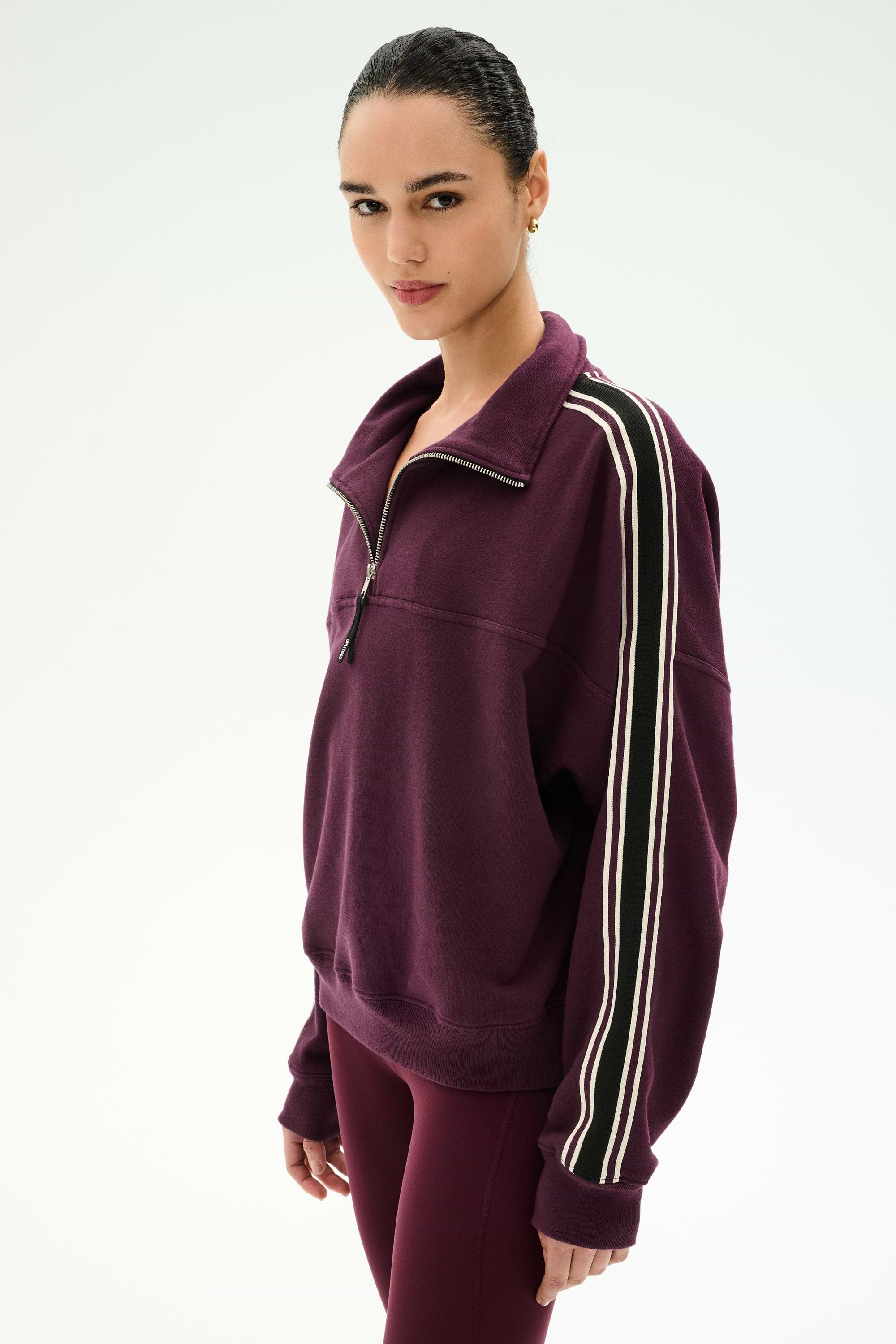 A person is wearing the SPLITS59 Goldie French Terry Oversized Half Zip in Dark Cherry/Black, featuring white-striped sleeves and matching leggings, crafted from soft cotton fabric, while standing against a plain light background.
