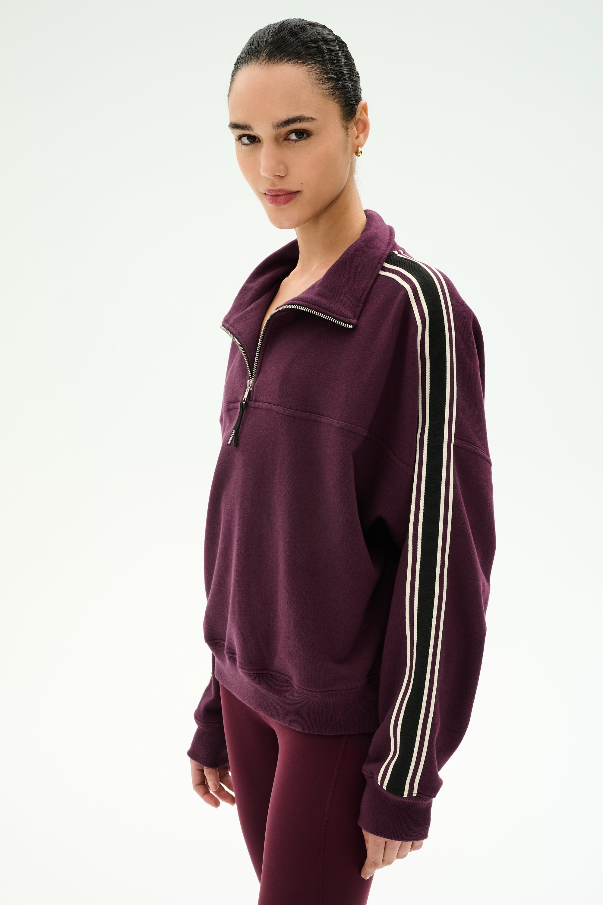 A person is wearing the SPLITS59 Goldie French Terry Oversized Half Zip in Dark Cherry/Black, featuring white-striped sleeves and matching leggings, crafted from soft cotton fabric, while standing against a plain light background.
