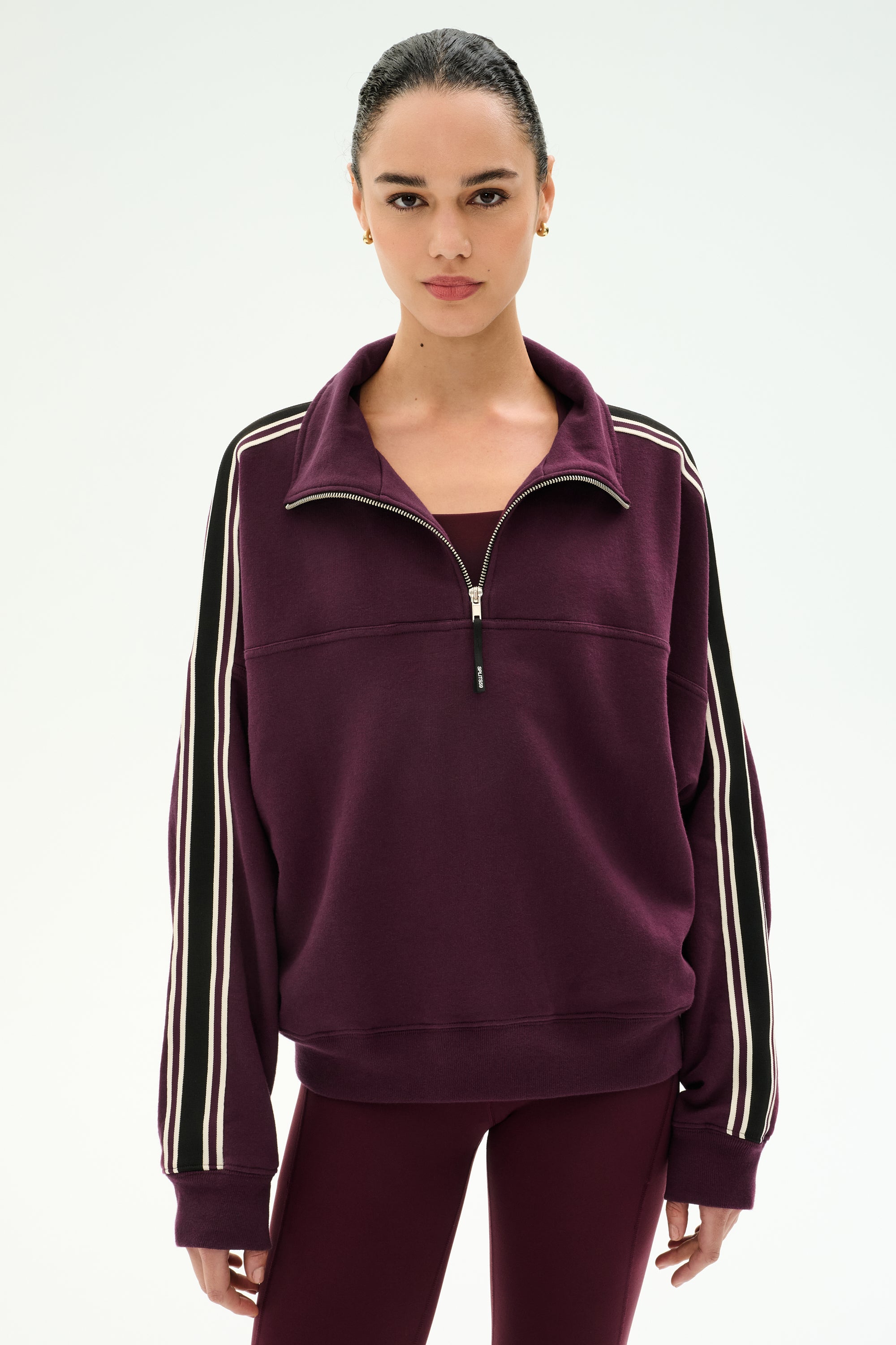 A woman wearing the SPLITS59 Goldie French Terry Oversized Half Zip in Dark Cherry/Black, crafted from soft cotton-blend fabric with white striped sleeves, paired with matching maroon leggings, stands against a plain light background.