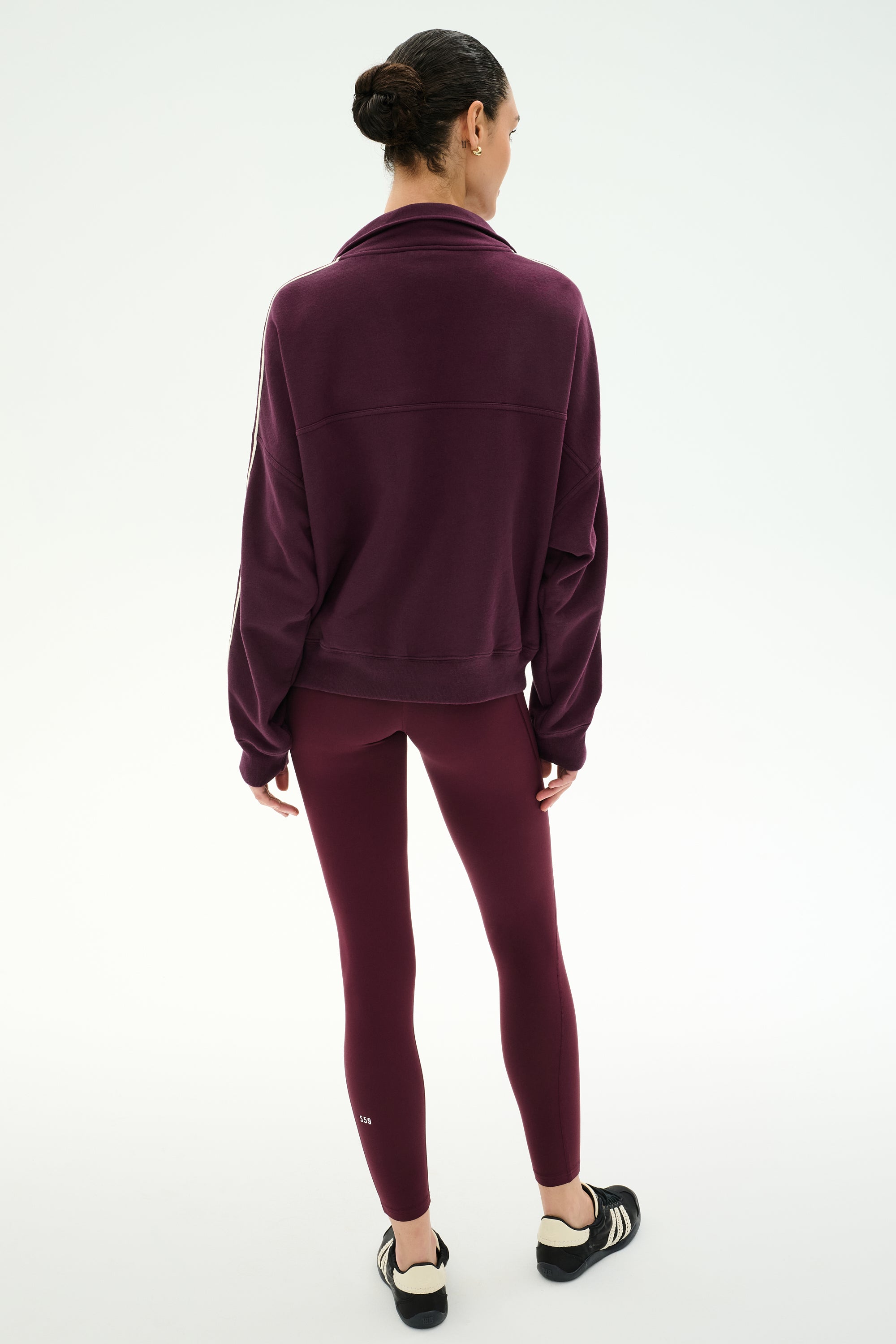 A person stands with their back to the camera on a plain white background, wearing the SPLITS59 Goldie French Terry Oversized Half Zip in Dark Cherry/Black, styled with matching maroon leggings and black slip-on shoes.