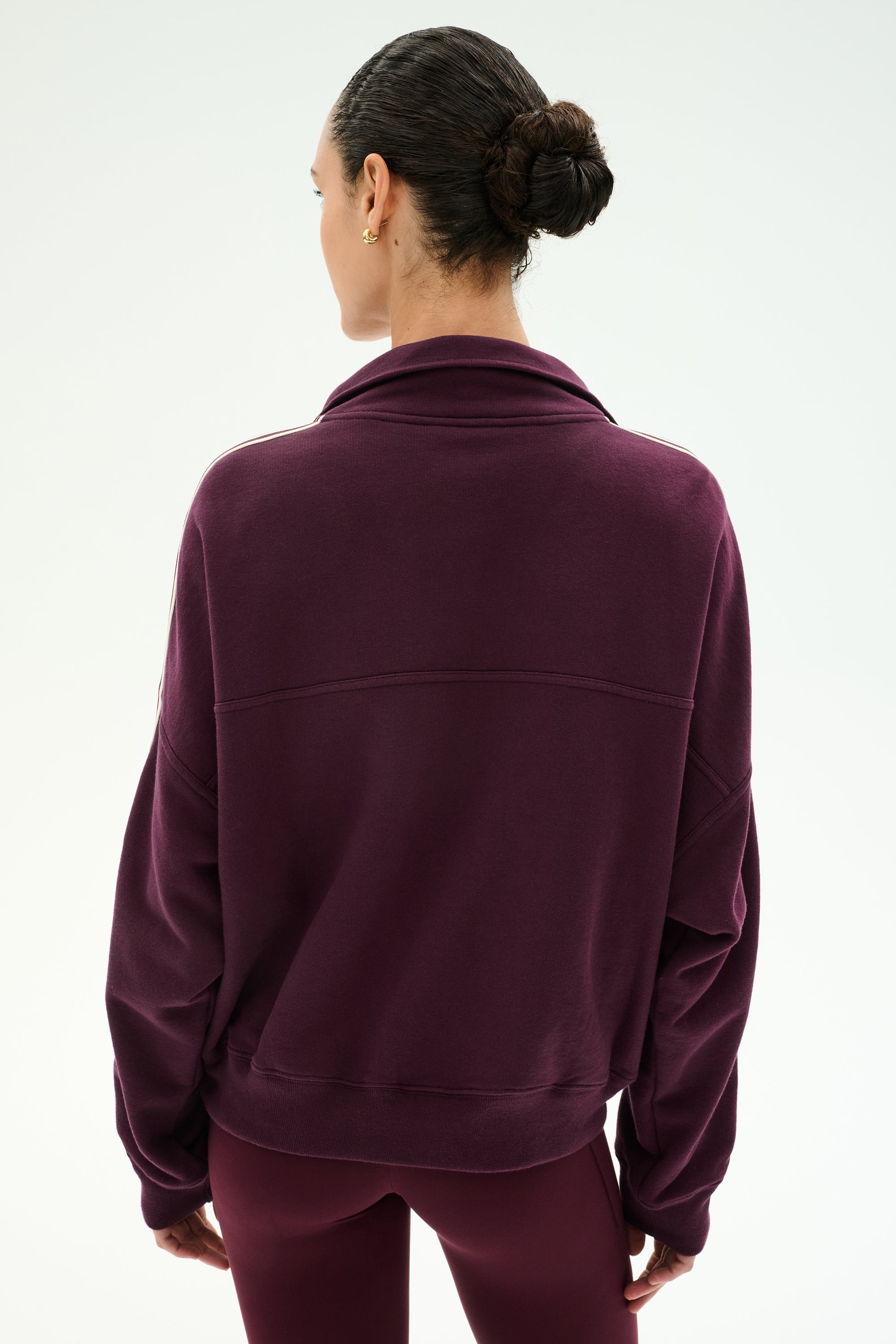 A woman with dark hair in a bun stands with her back to the camera against a plain light background, wearing the SPLITS59 Goldie French Terry Oversized Half Zip in Dark Cherry/Black.