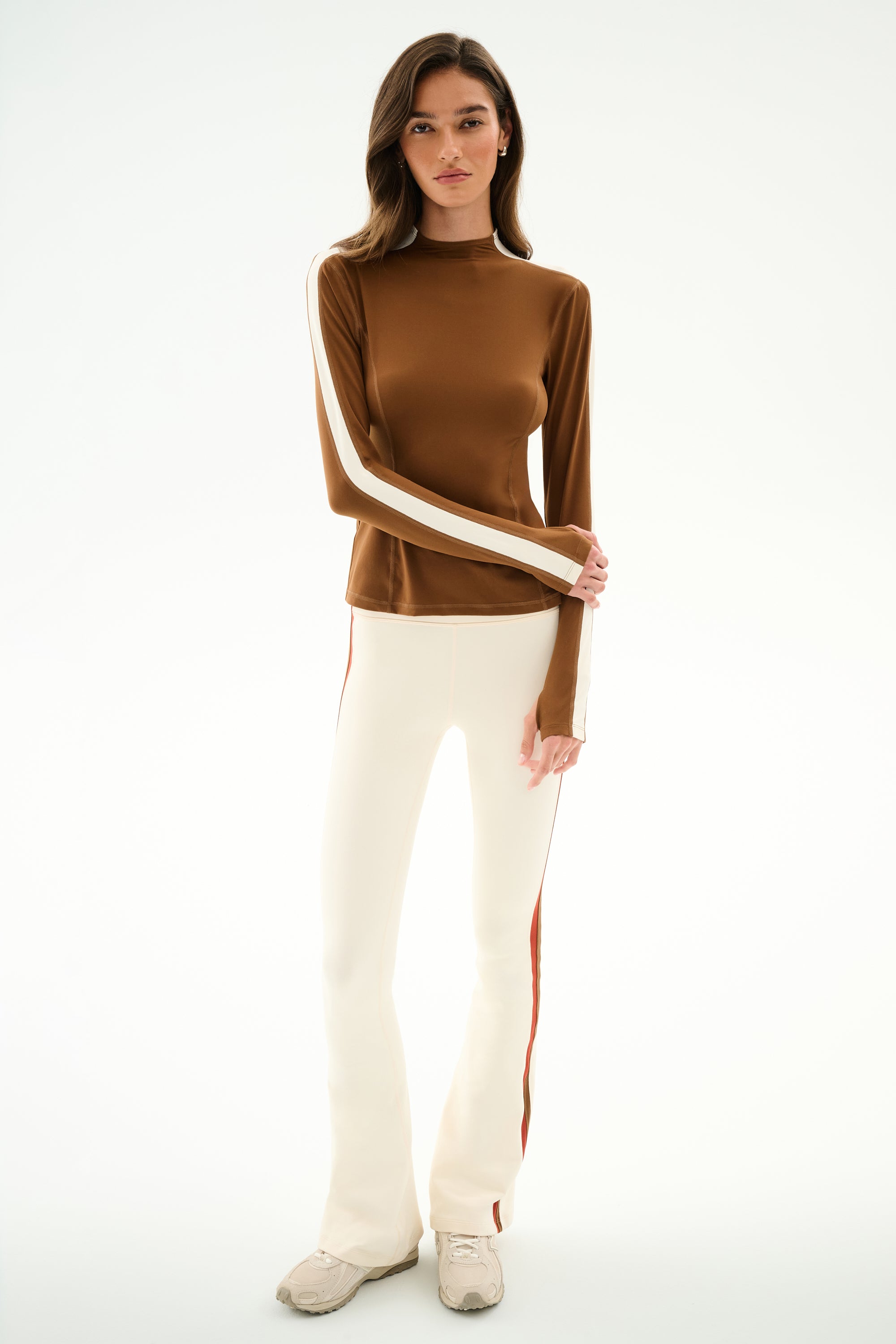 A person faces forward wearing the SPLITS59 Isabelle Airweight Lite Mock Neck - Toffee/Creme, cream flared spandex pants with side stripes, and white sneakers.
