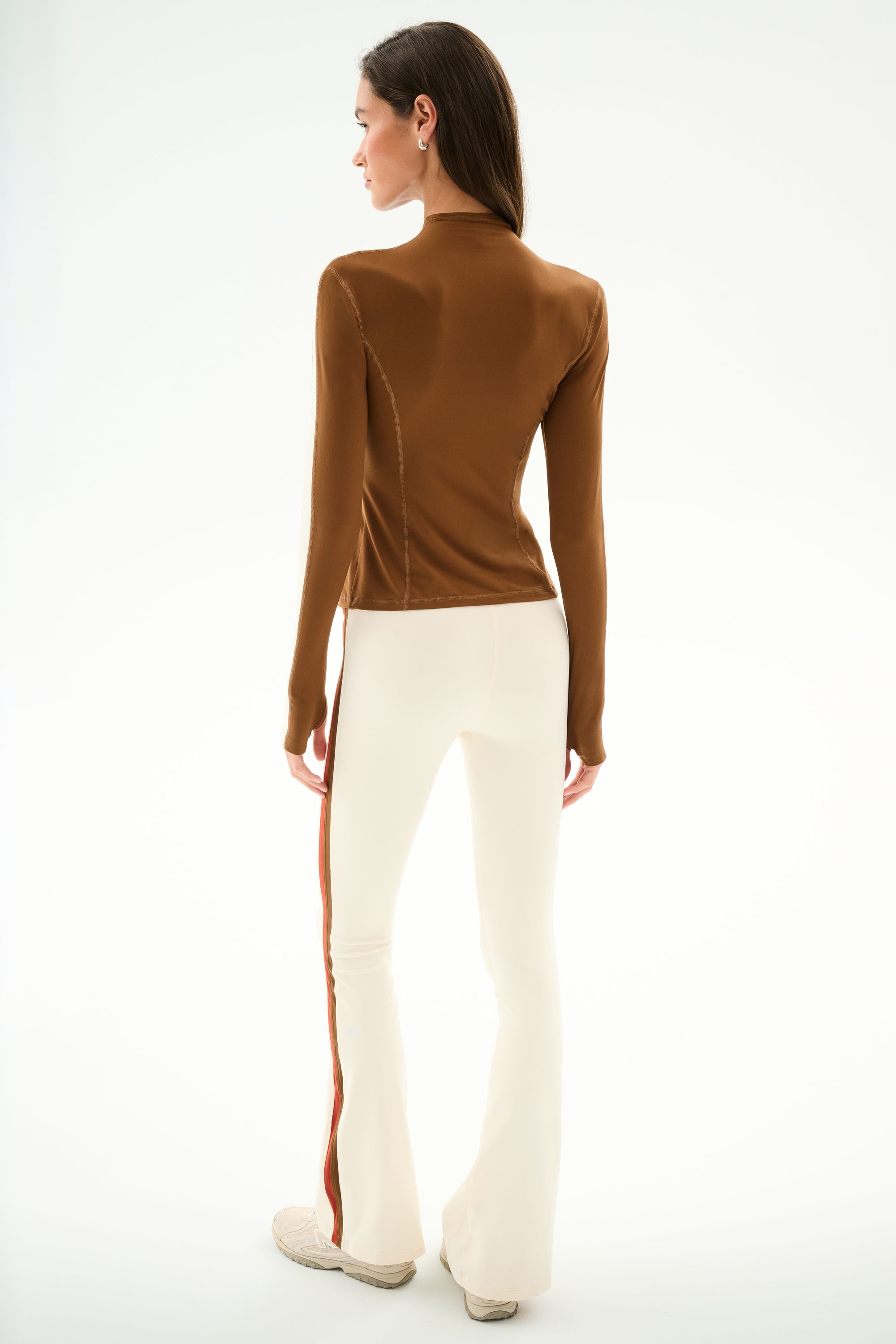 A person wears the Isabelle Airweight Lite Mock Neck - Toffee/Creme by SPLITS59, styled with white flared pants featuring a red side stripe, against a plain white background.