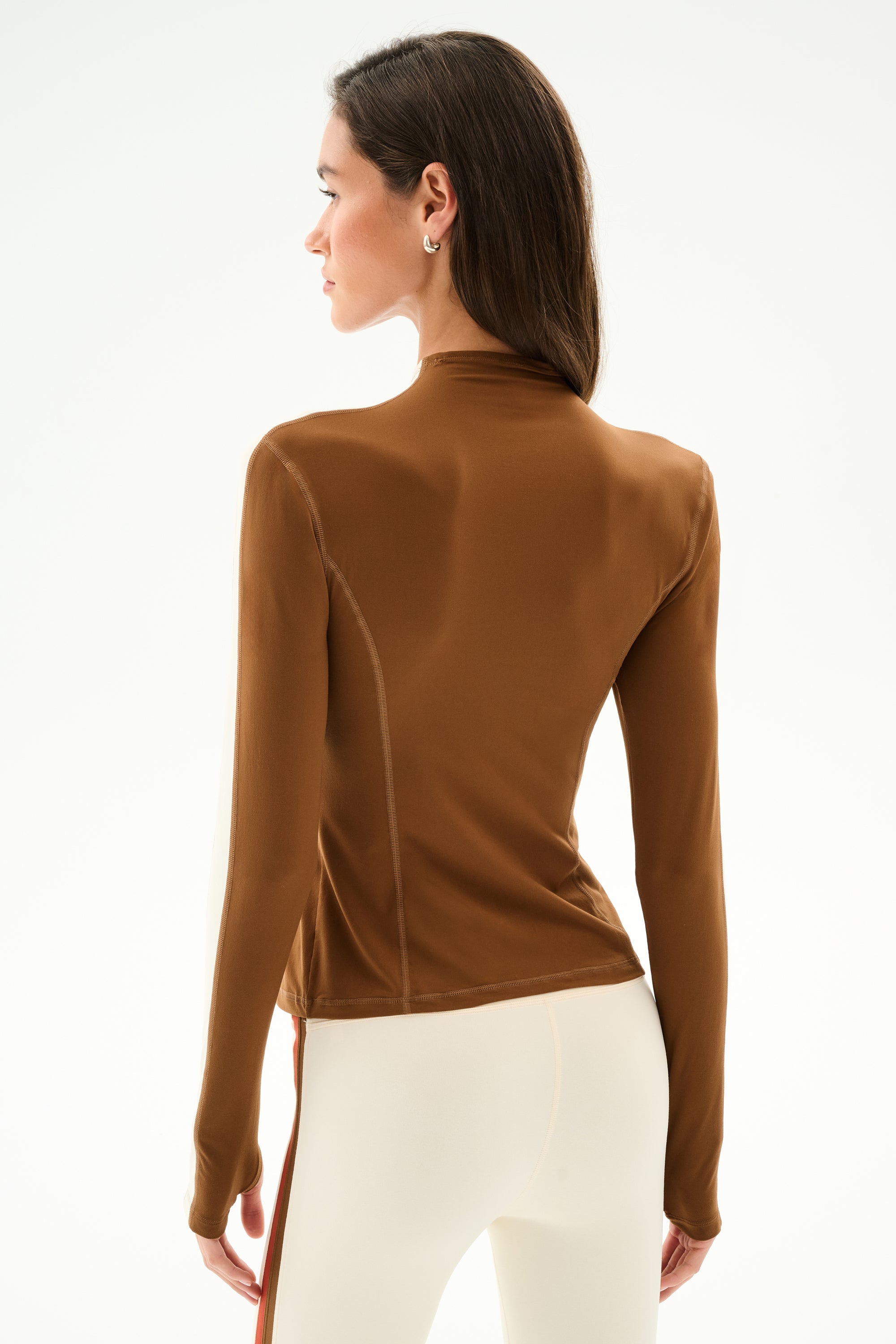 A woman faces away, wearing the SPLITS59 Isabelle Airweight Lite Mock Neck - Toffee/Creme, paired with off-white pants with a side stripe, against a plain white background.