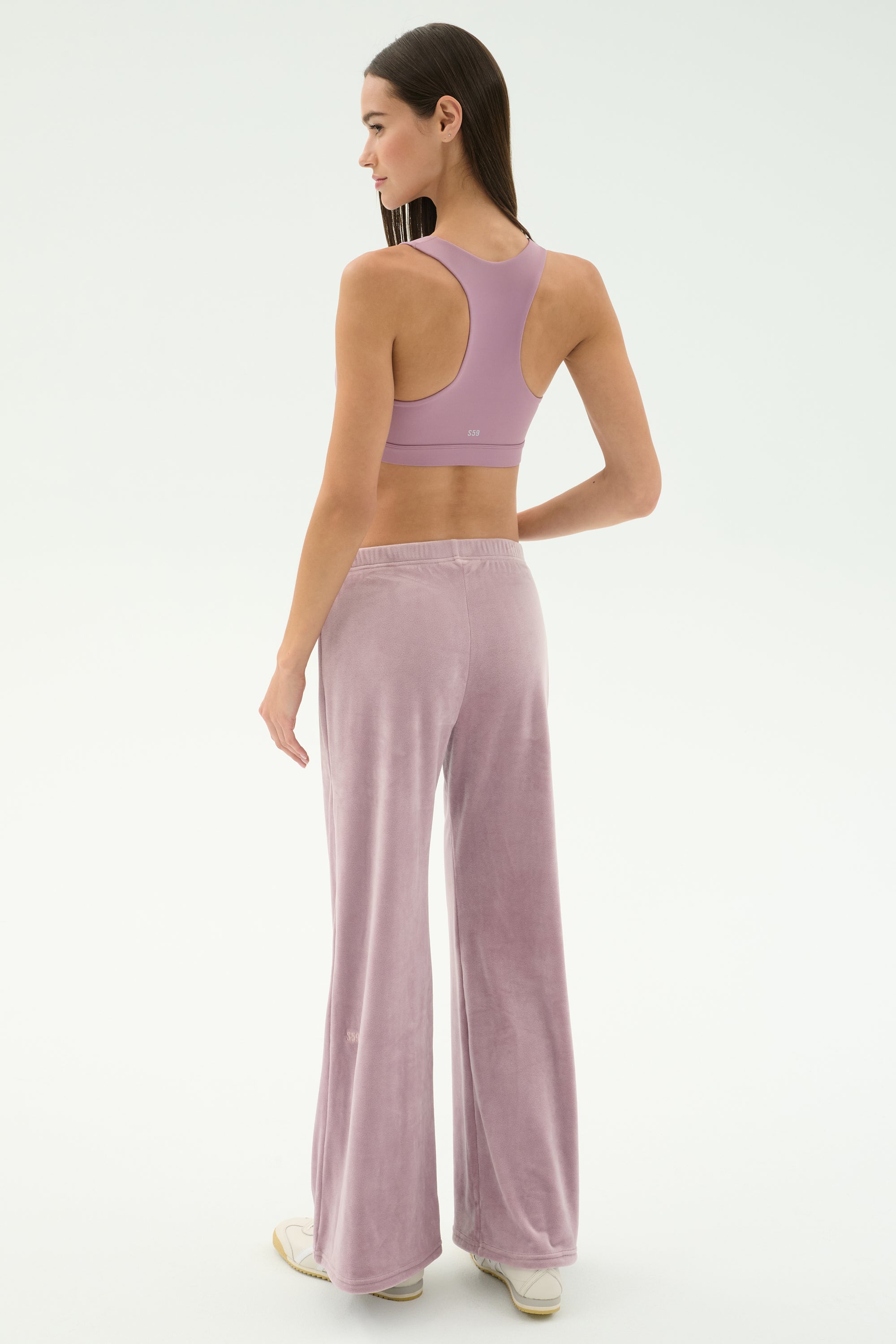 A woman stands in a studio facing away, wearing SPLITS59’s Joy Velour Pant in Blush and a matching mauve sports bra, set against a plain white background.