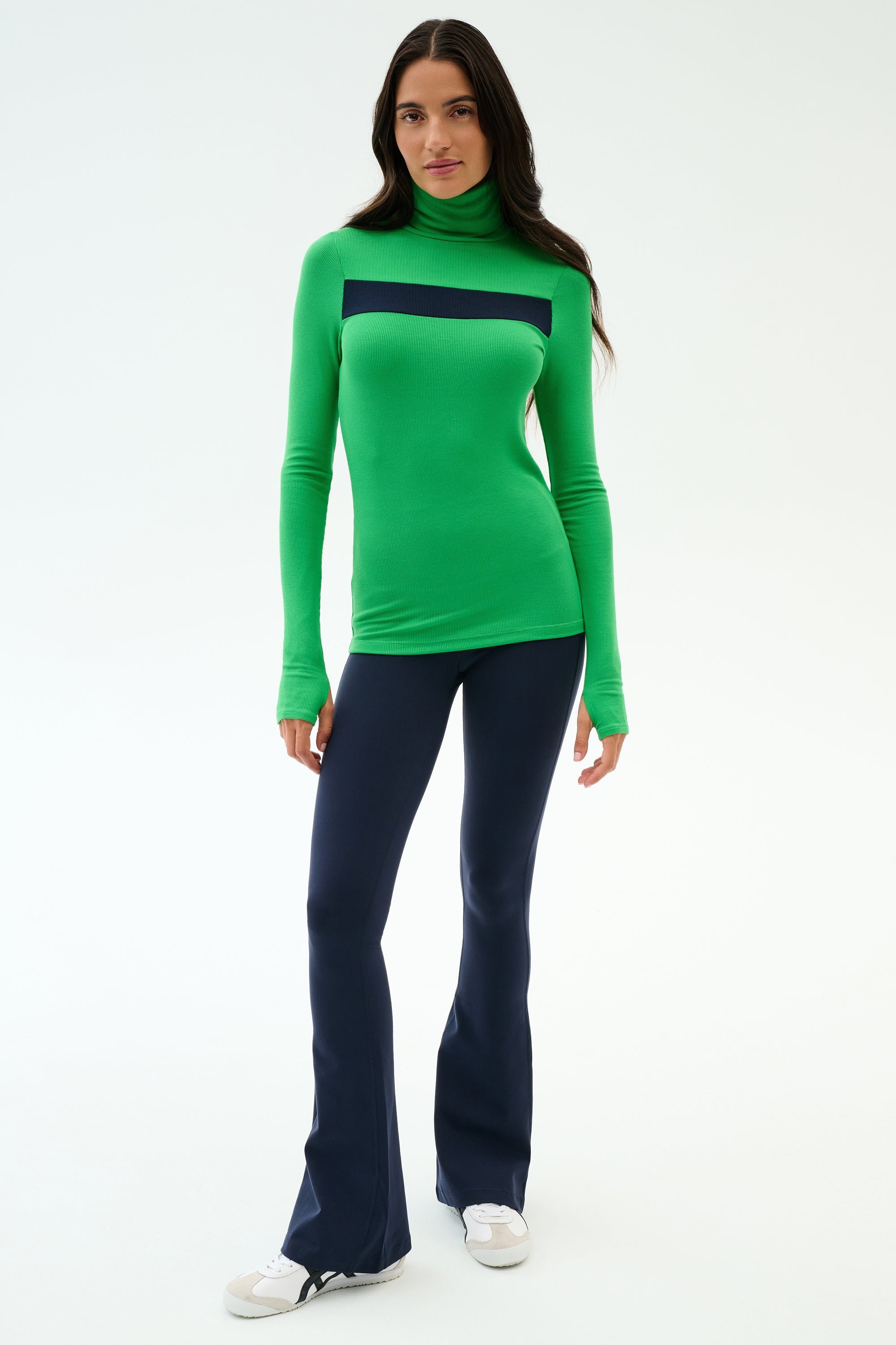 A woman faces forward wearing the SPLITS59 Lennon Rib Turtleneck in Grass/Indigo and navy flared pants with white sneakers—ideal for yoga or errands—against a plain white background.