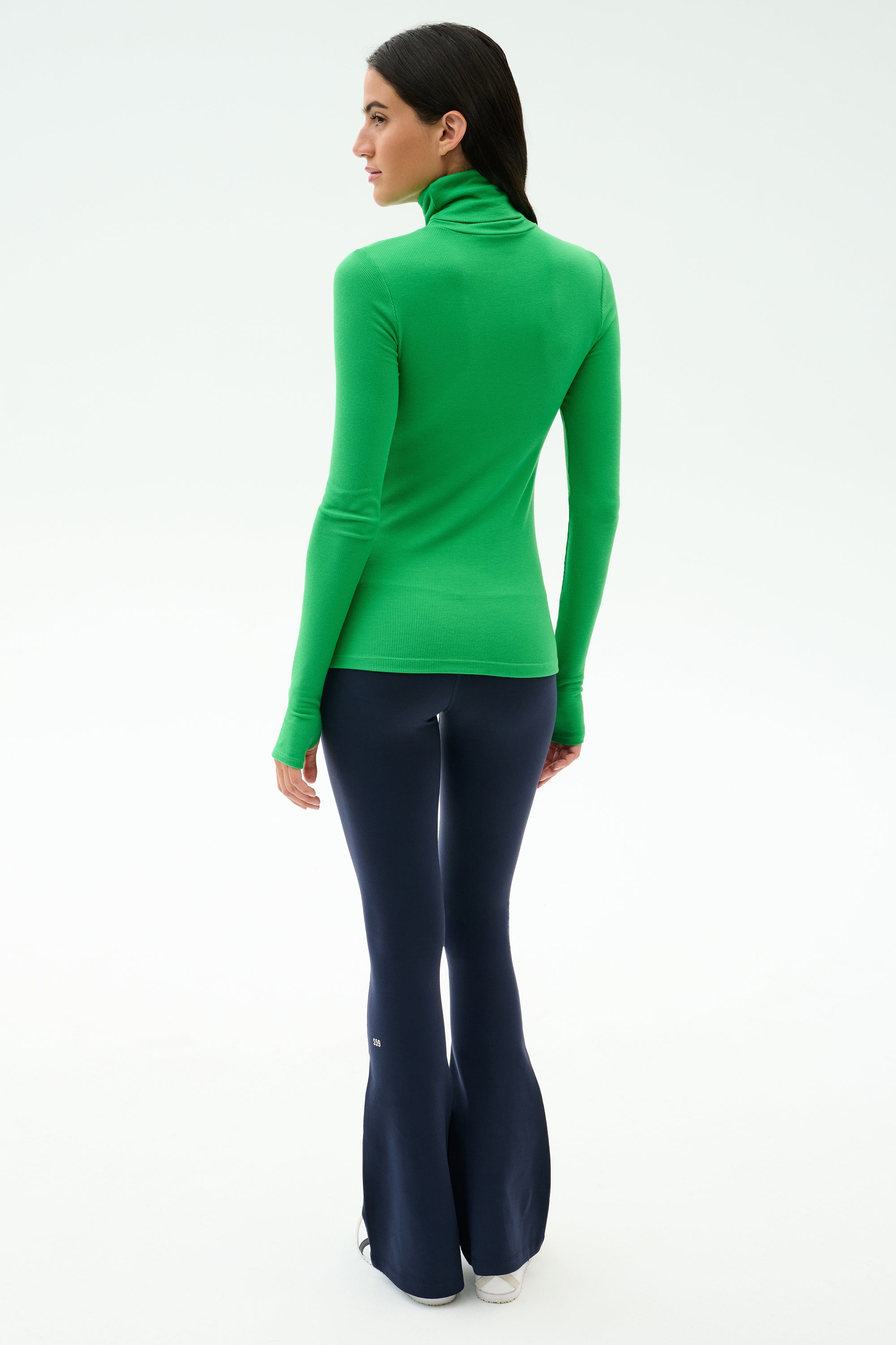 A person stands facing away, wearing the SPLITS59 Lennon Rib Turtleneck in Grass/Indigo and dark flared pants against a plain, light background.