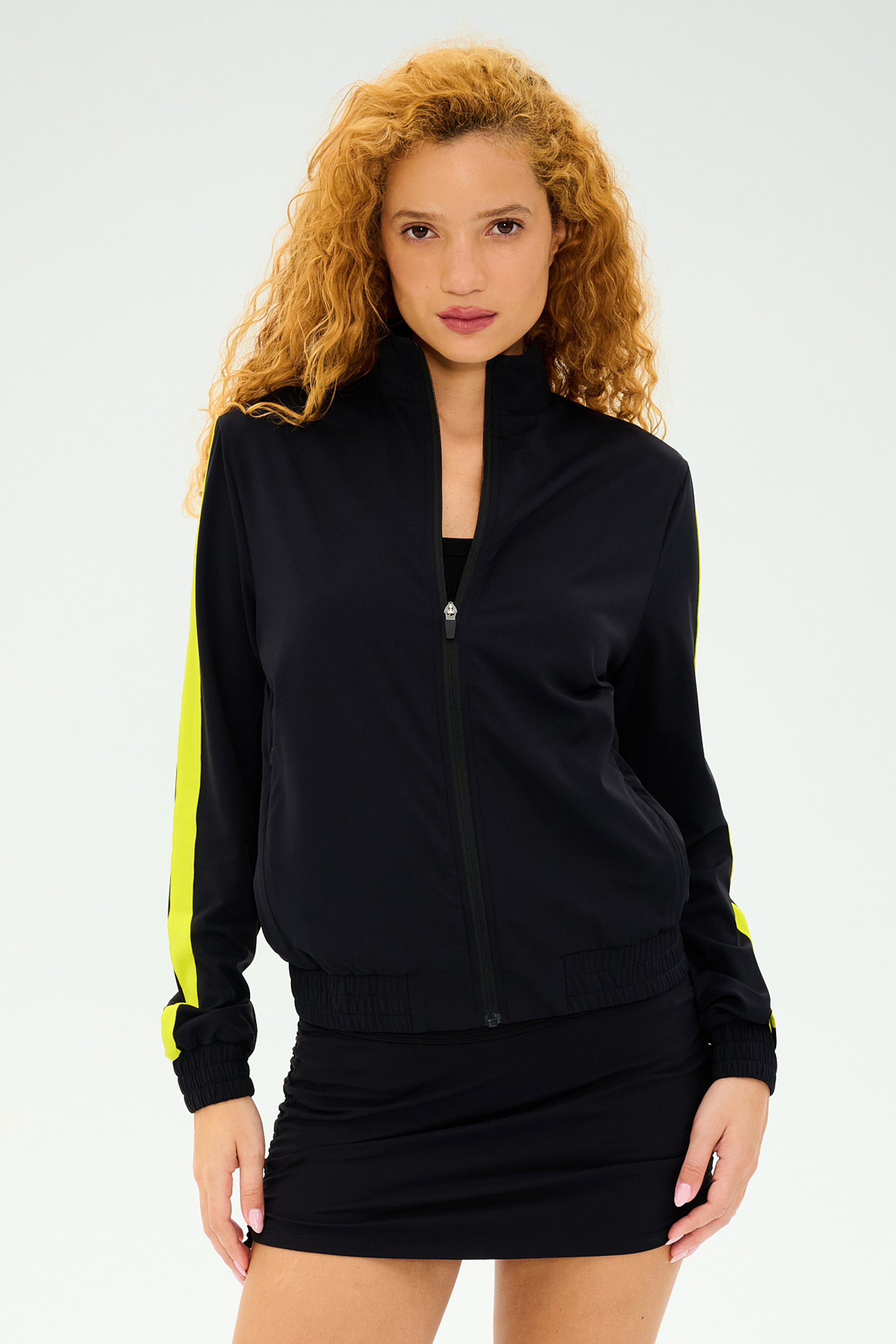 Against a plain background, a person with curly hair wears the SPLITS59 Max Rigor Track Jacket in Black/Chartreuse and a black skirt, highlighting its sleek design reminiscent of Max Rigor Track Pant's renowned style.
