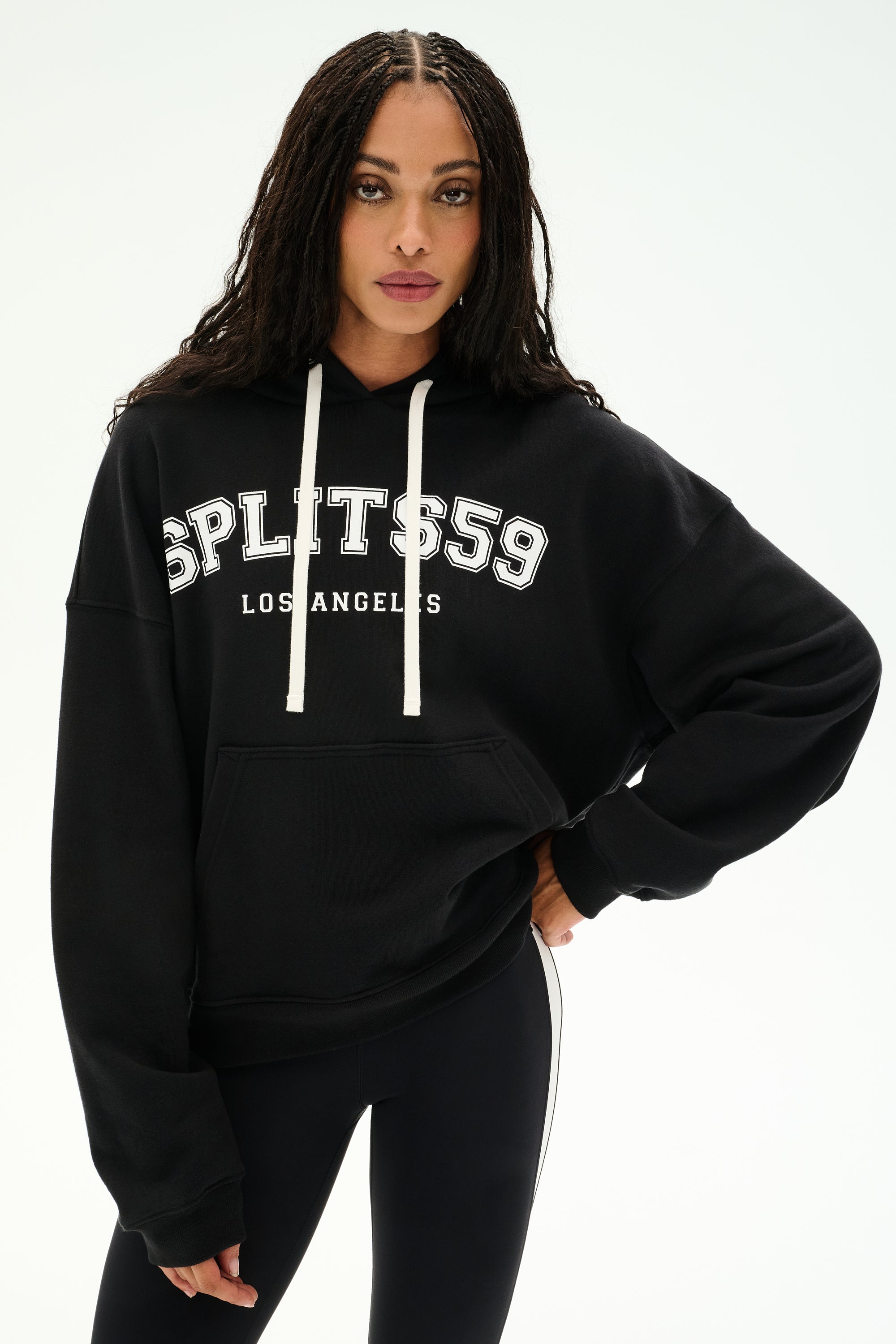 A woman is wearing the SPLITS59 Stella French Terry Oversized Hoodie in black with "Splits59 Los Angeles" text, paired with black leggings featuring white side stripes and white drawstrings, as she stands against a plain white background.